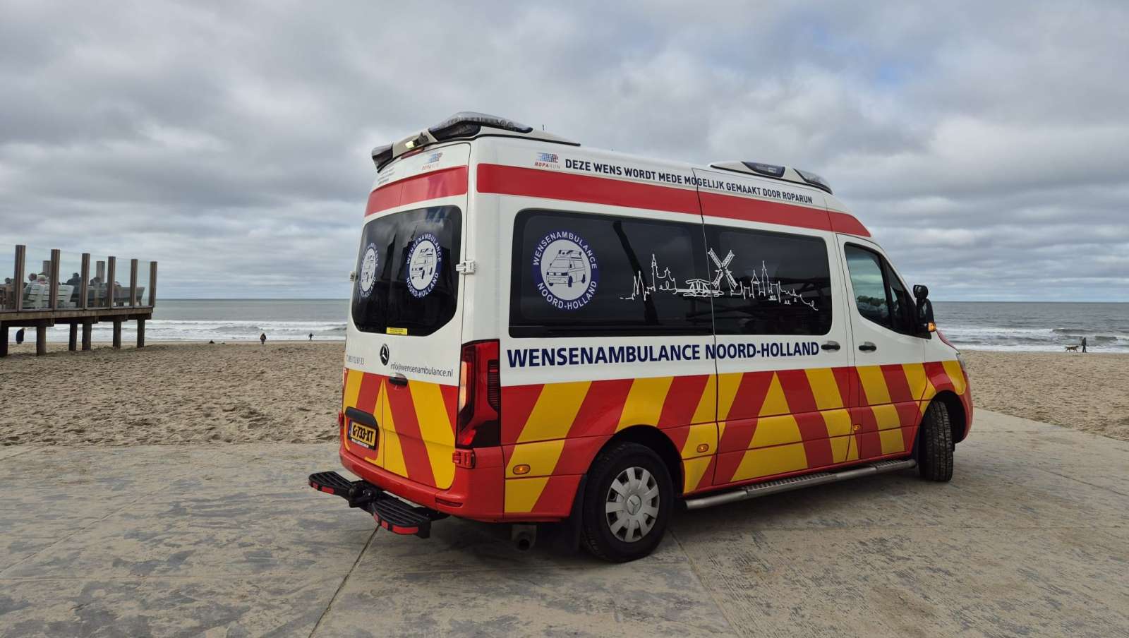 Een Wensambulance Noord-Holland geparkeerd bij een strand met deels bewolkte hemel op de achtergrond.