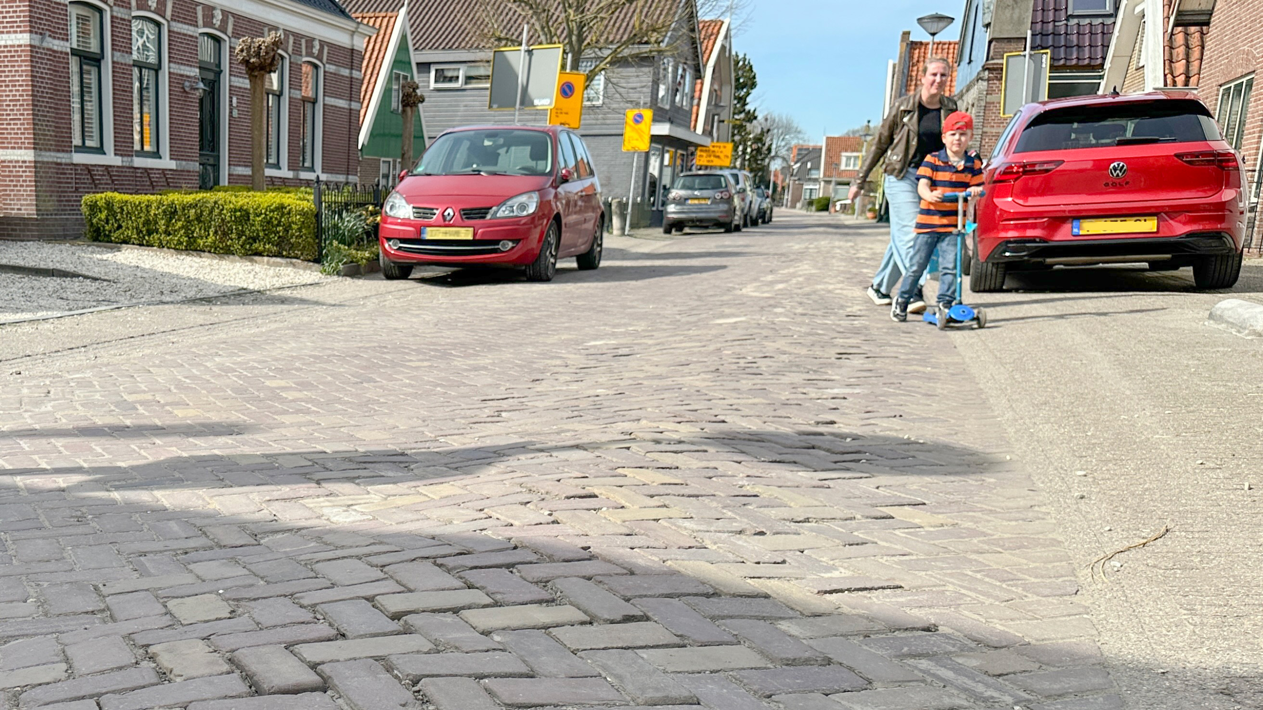 Een kind rijdt op een blauwe step naast een volwassene op een rustige straat met geparkeerde auto's en huizen.
