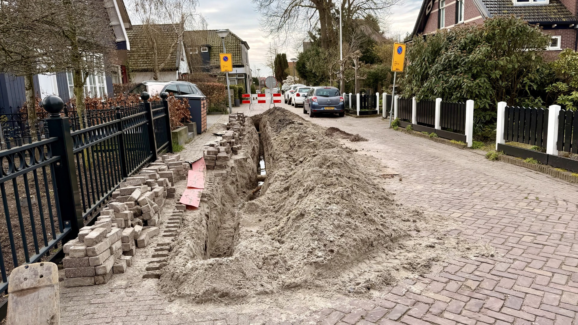 Geopende straat in een woonwijk met een geul gevuld met zand en opgebroken stoeptegels aan de zijkant.