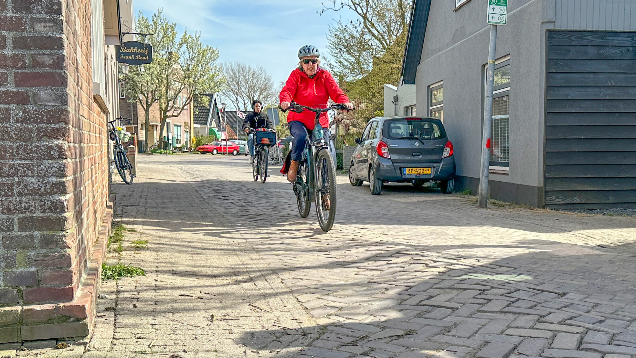 Twee personen fietsen door een rustige straat, met een bord van een bakkerij op de achtergrond.