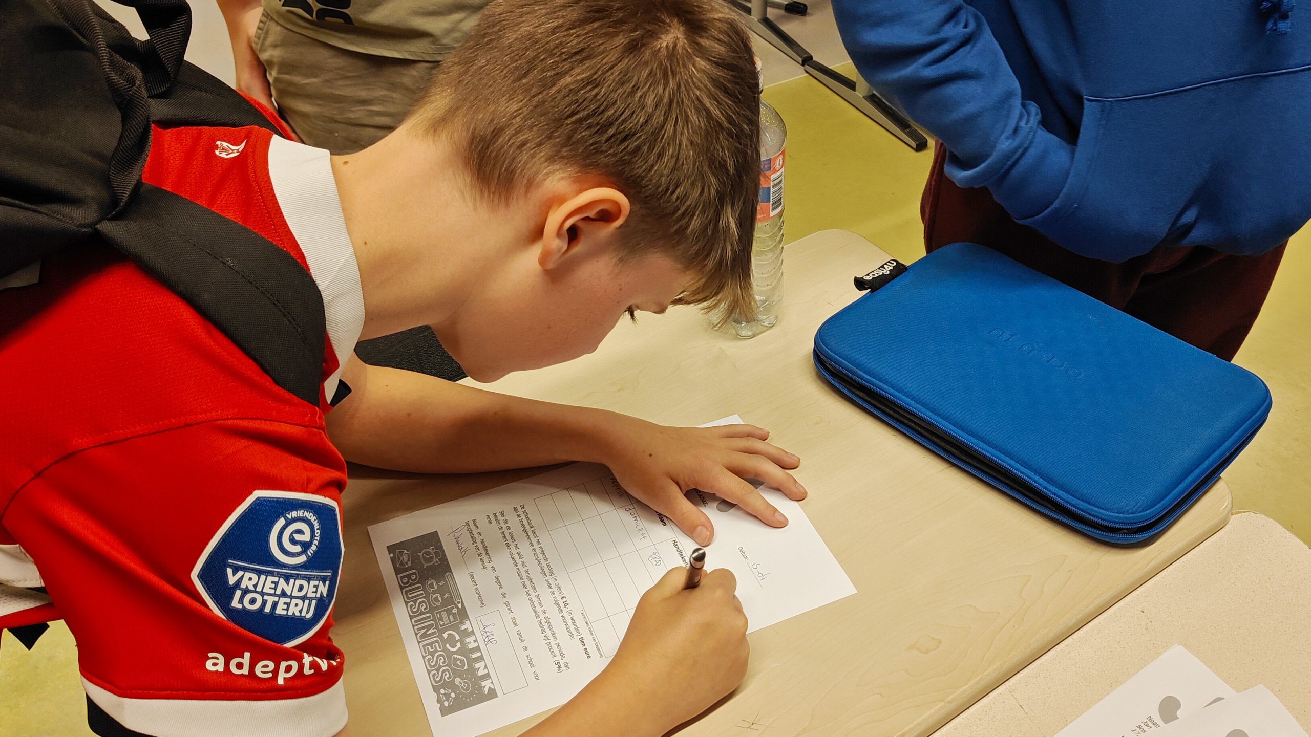Een jongen in een rode trui met rugzak ondertekent een formulier aan tafel, naast een blauwe hoes en een flesje water.