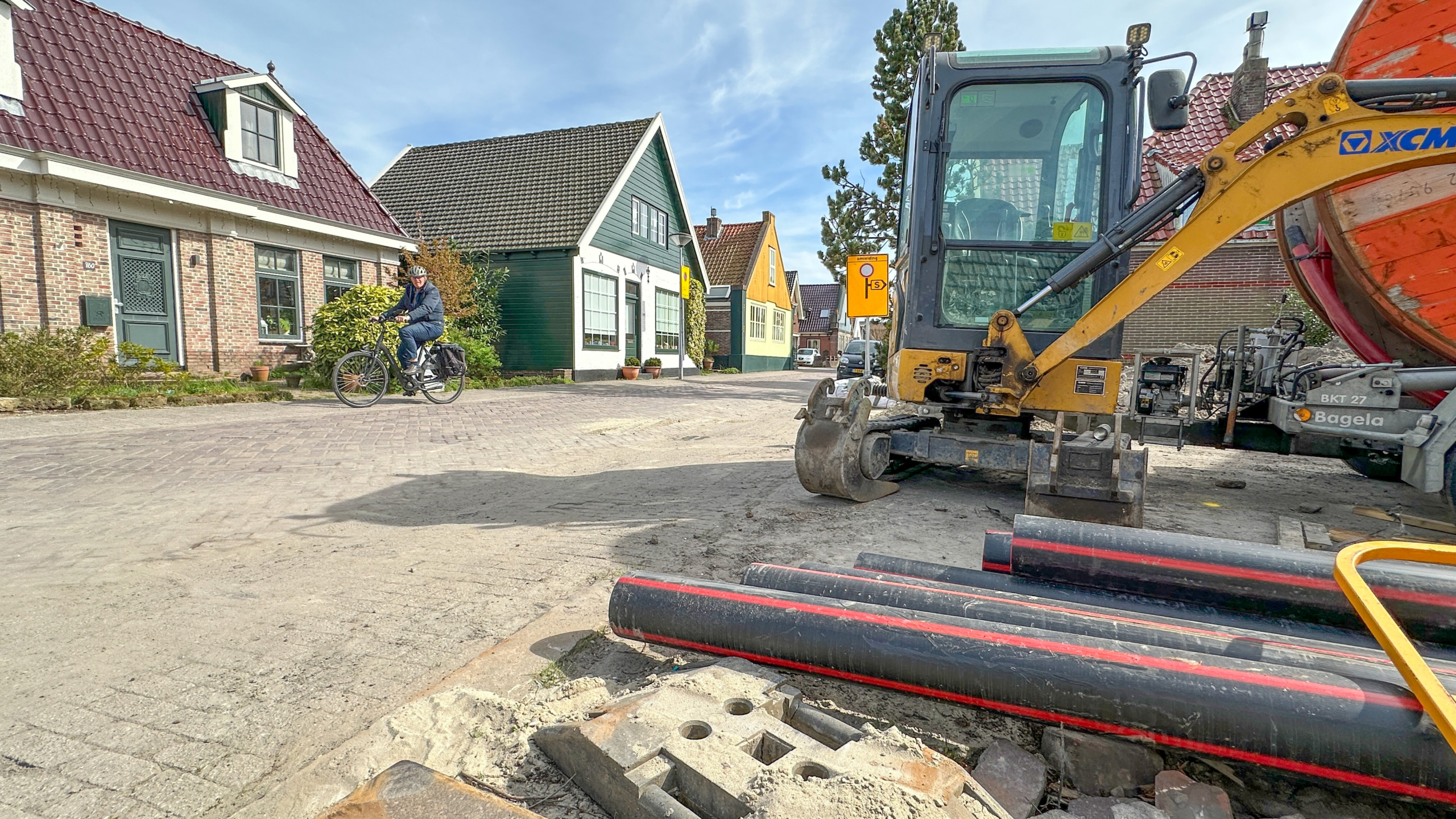 Een fietser rijdt langs een bouwplaats met een graafmachine en buizen in een straat met traditionele huizen.