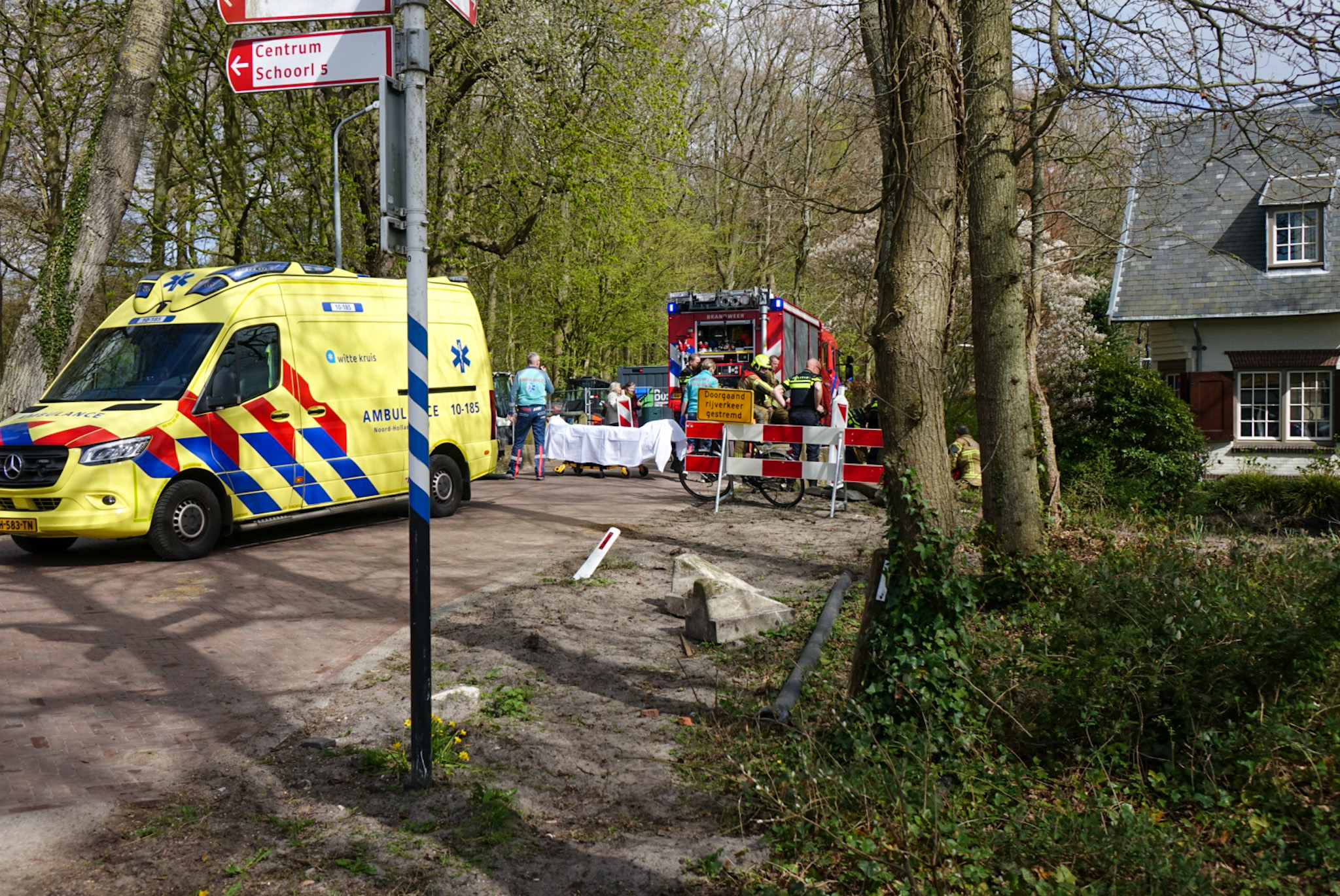 Gele ambulance en rode brandweerwagen bij wegafzetting in bosrijke omgeving; hulpdiensten aanwezig.
