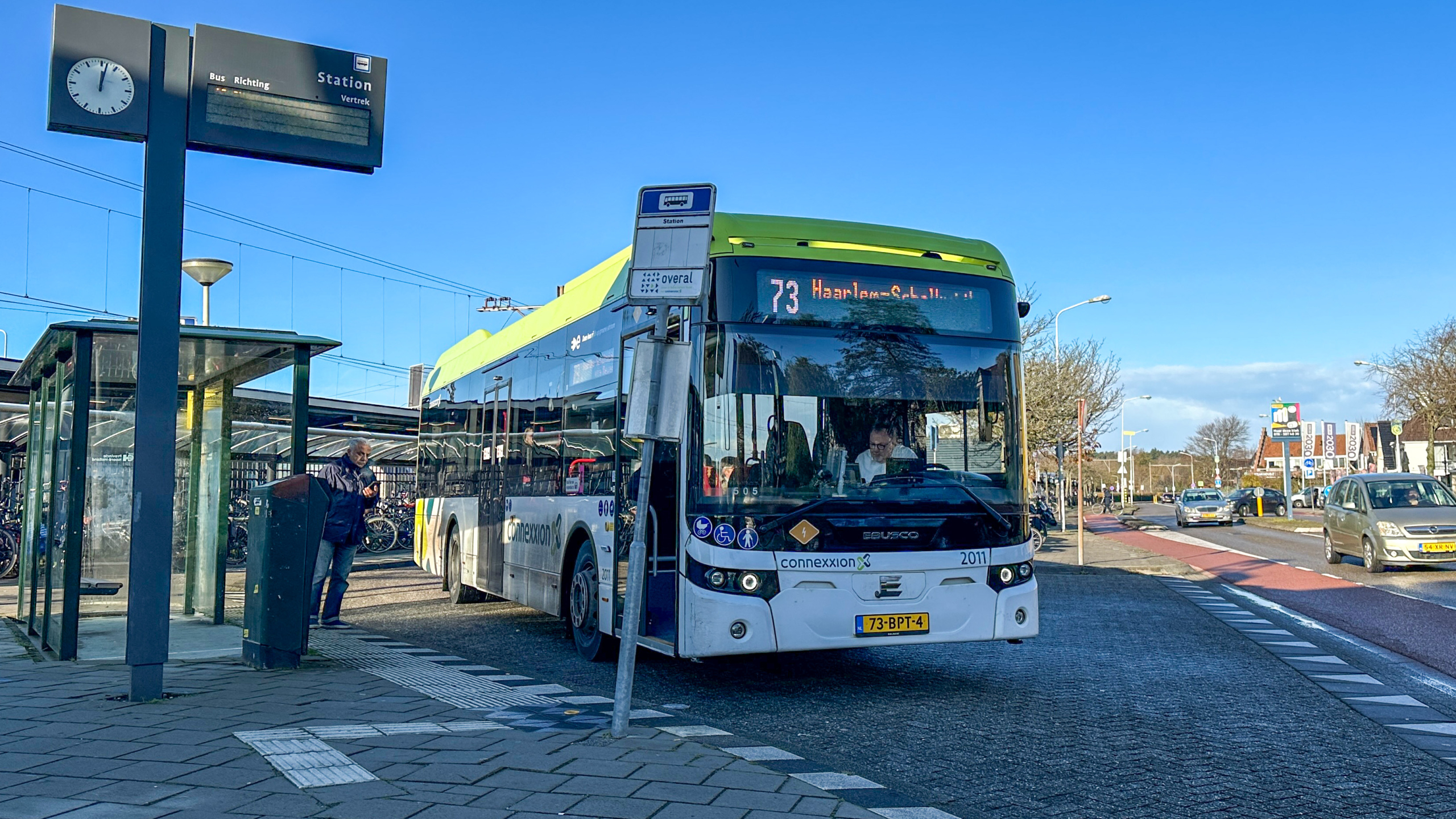 Bus 73 naar Haarlem overdag bij een bushalte, met een busstation in de achtergrond en een voorbijganger.