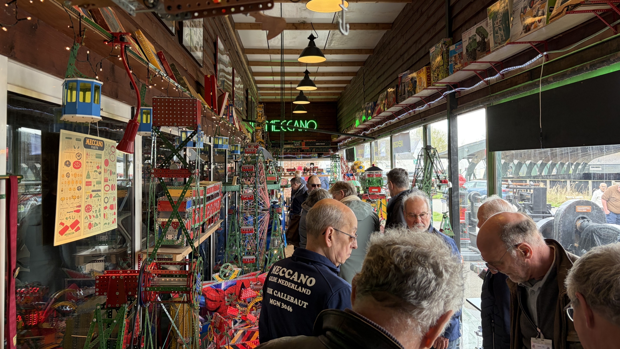 Mensen verzamelen zich in een winkel met Meccano displays en bordspellen aan de muren, terwijl ze verschillende modellen bekijken.