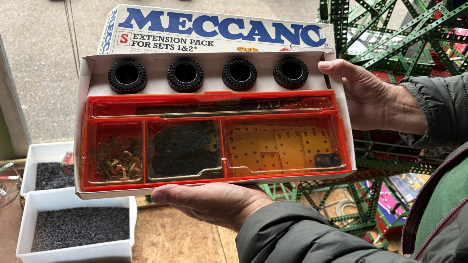 Een persoon houdt een Meccano uitbreidingsset vast met verschillende onderdelen, waaronder wielen en metalen platen.
