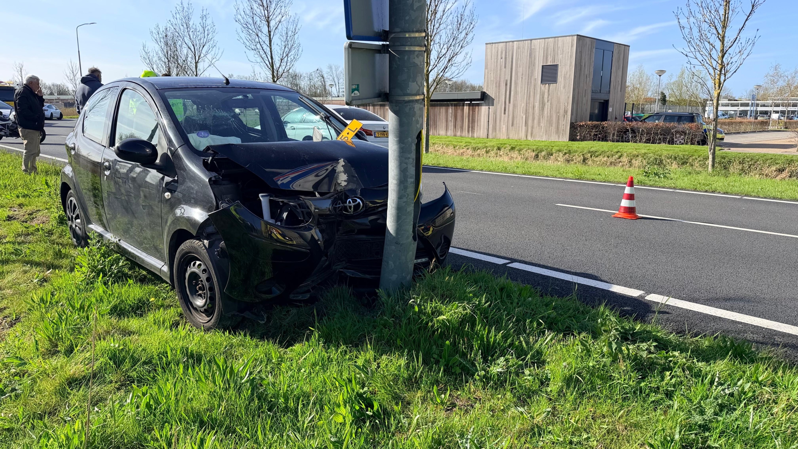 Auto met schade na botsing tegen een paal langs een weg, met verkeerskegel en mensen op de achtergrond.