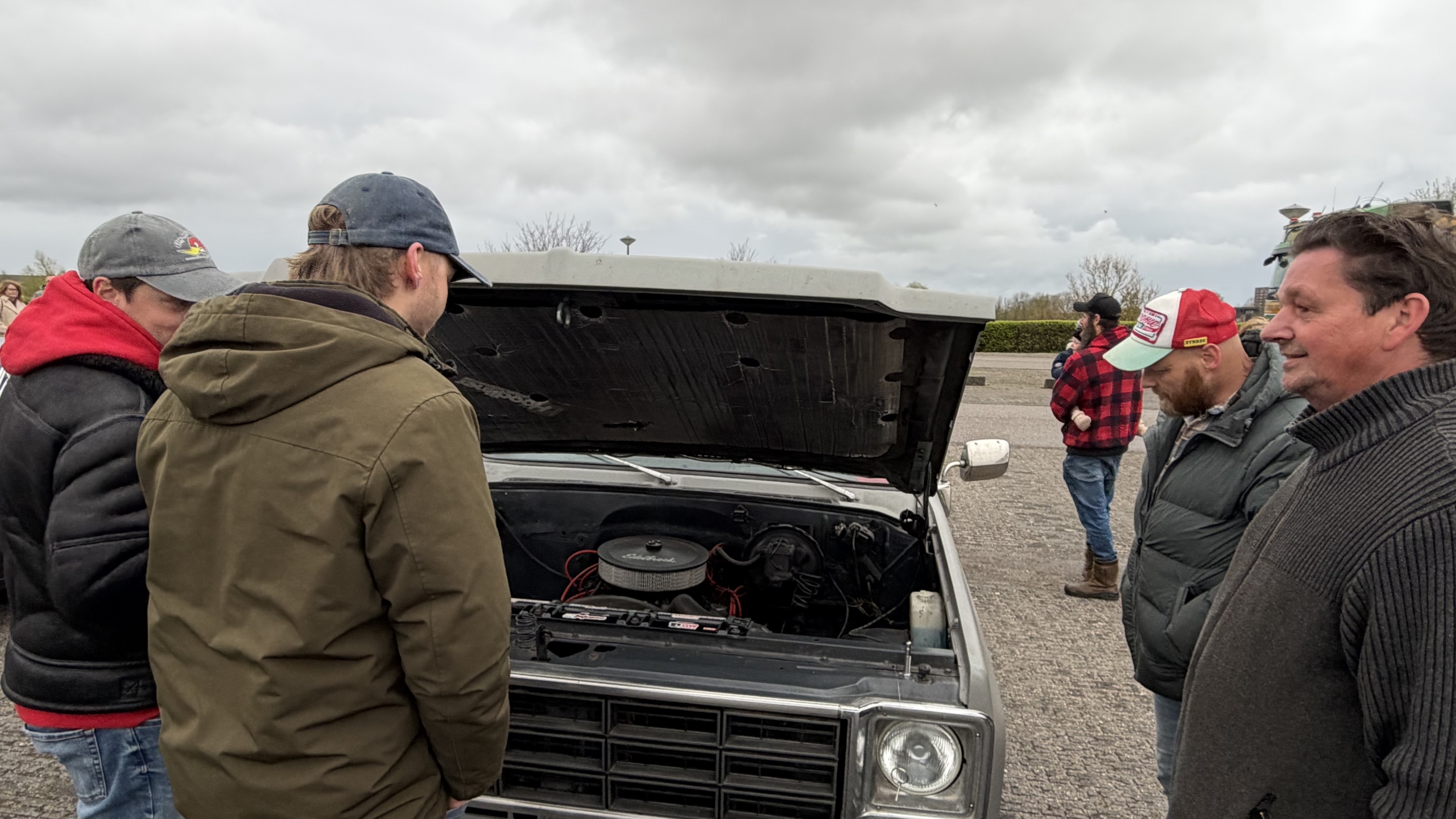 Mensen staan rond een auto met geopende motorkap en bekijken het motorblok op een parkeerplaats.