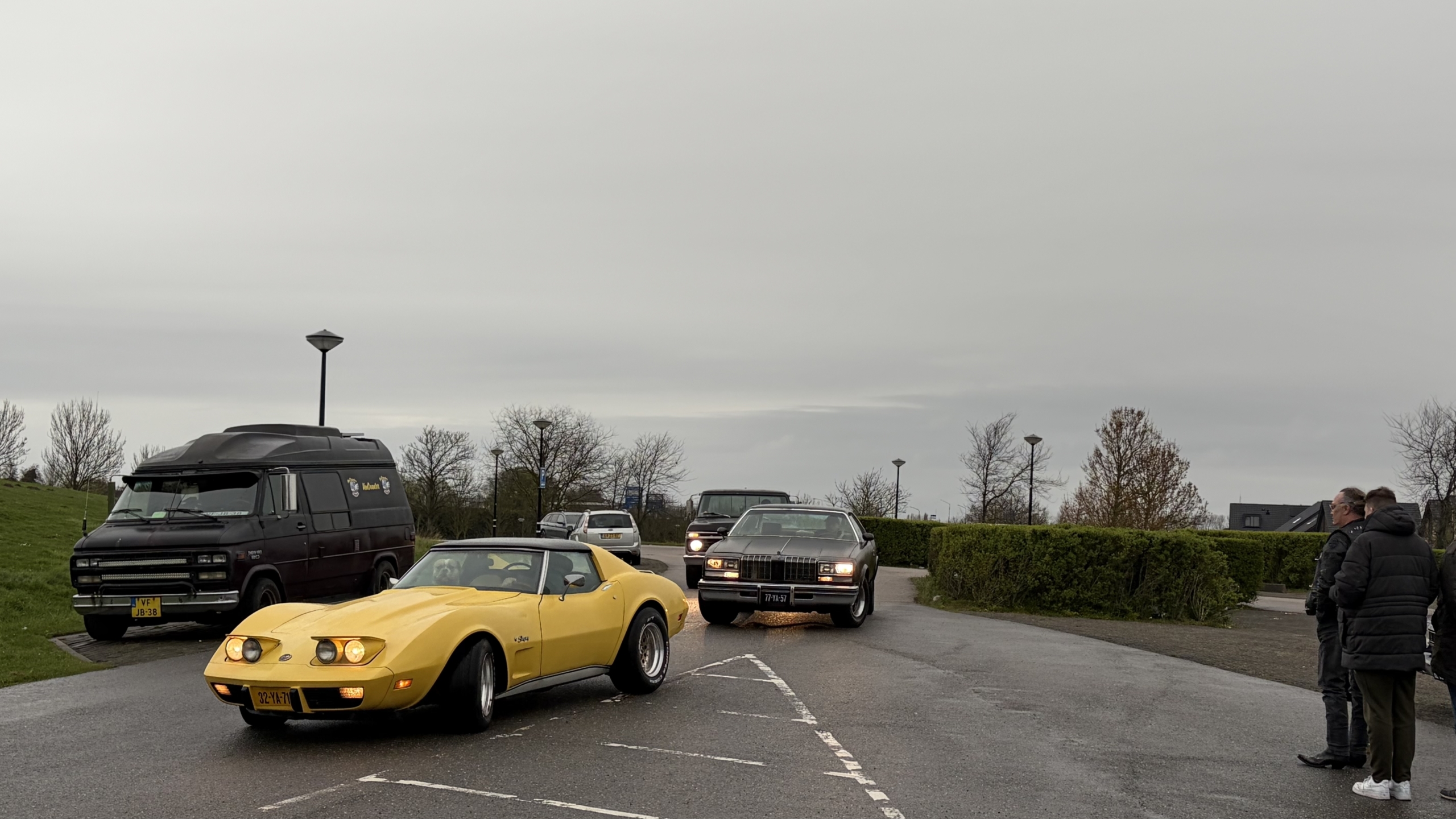 Gele sportauto en zwarte klassieke auto rijden op een weg terwijl drie personen toekijken.