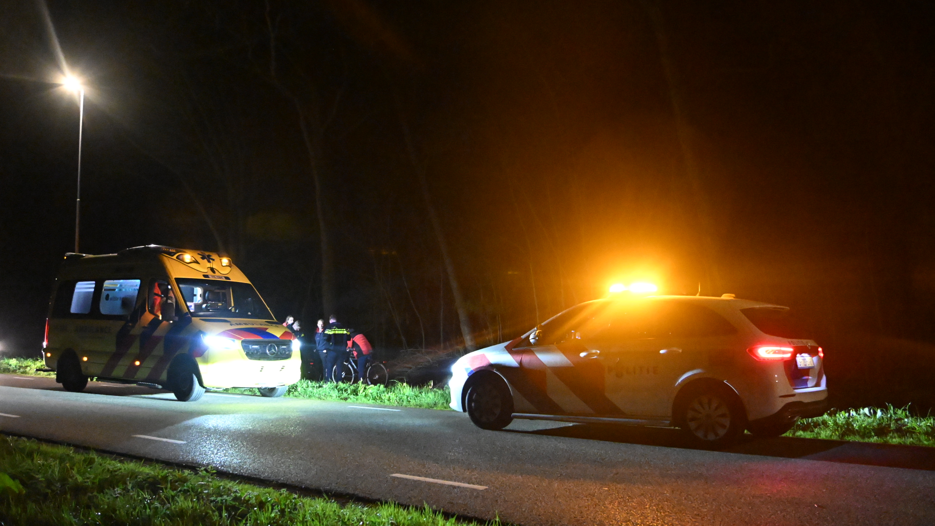 Een ambulance en een politieauto met zwaailichten staan 's nachts langs een weg, omgeven door mensen.