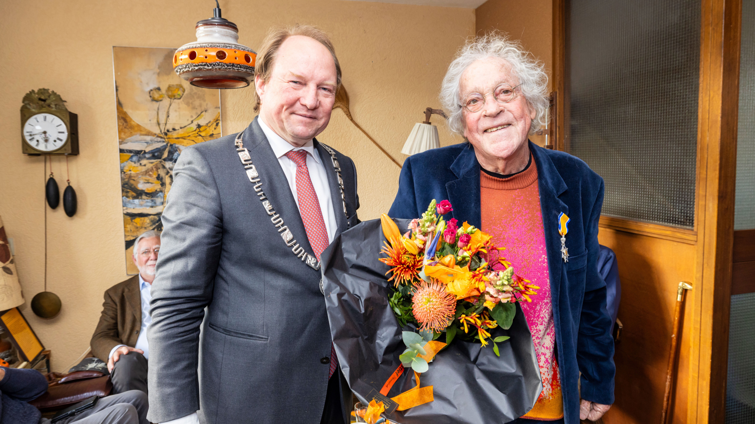 Twee mannen staan glimlachend naast elkaar, één met een bloemenboeket en een medaille.