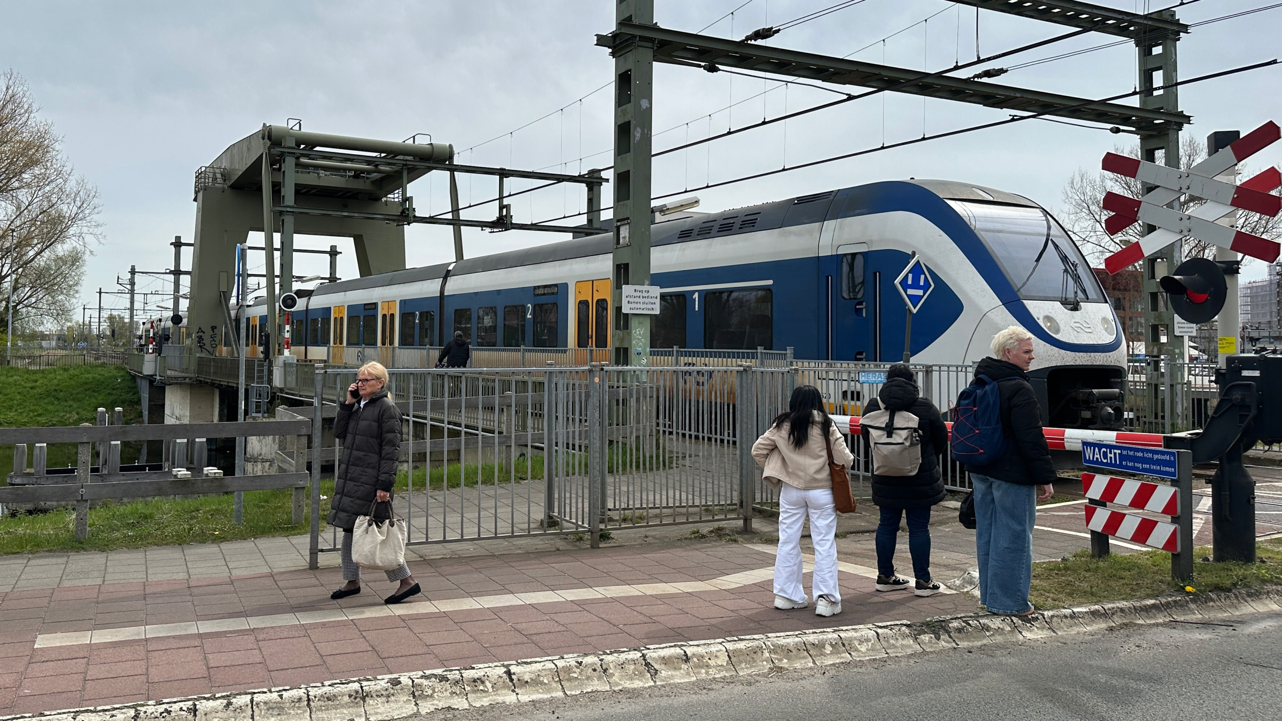 Een trein passeert een spoorwegovergang terwijl mensen op het trottoir wachten.