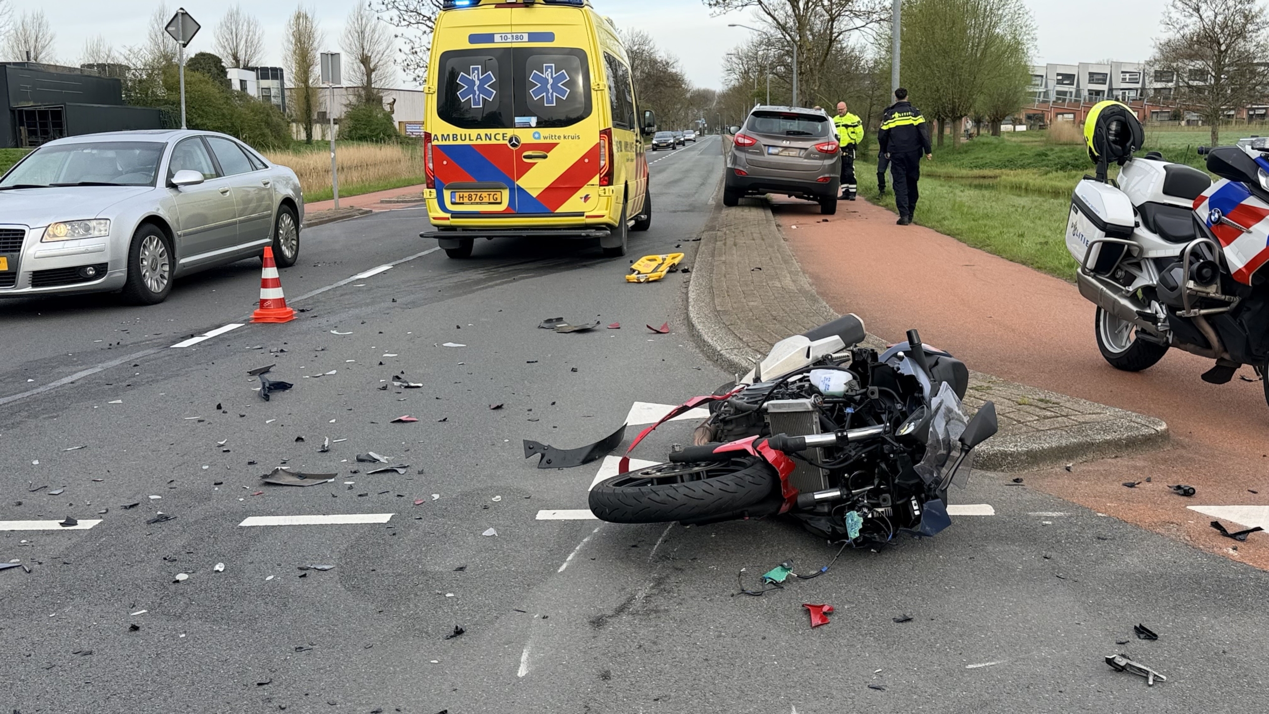 Verkeersongeluk met een omgevallen motorfiets, schade op de weg, en hulpverleningsvoertuigen, waaronder een ambulance en politiefiets.