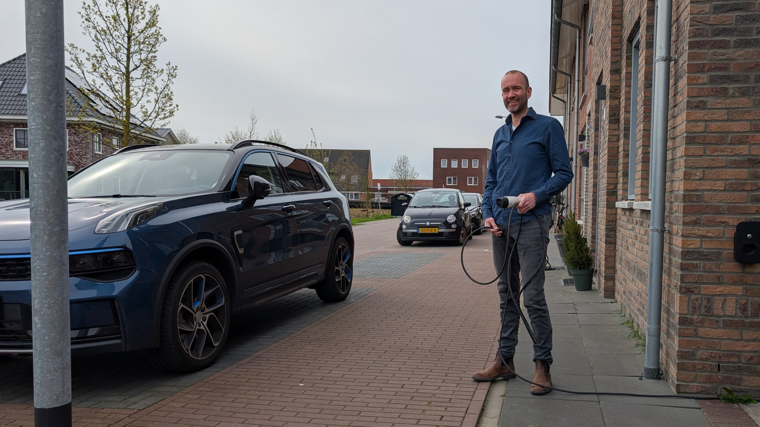 Man plugt een elektrische auto in op een straat met bakstenen huizen op de achtergrond.