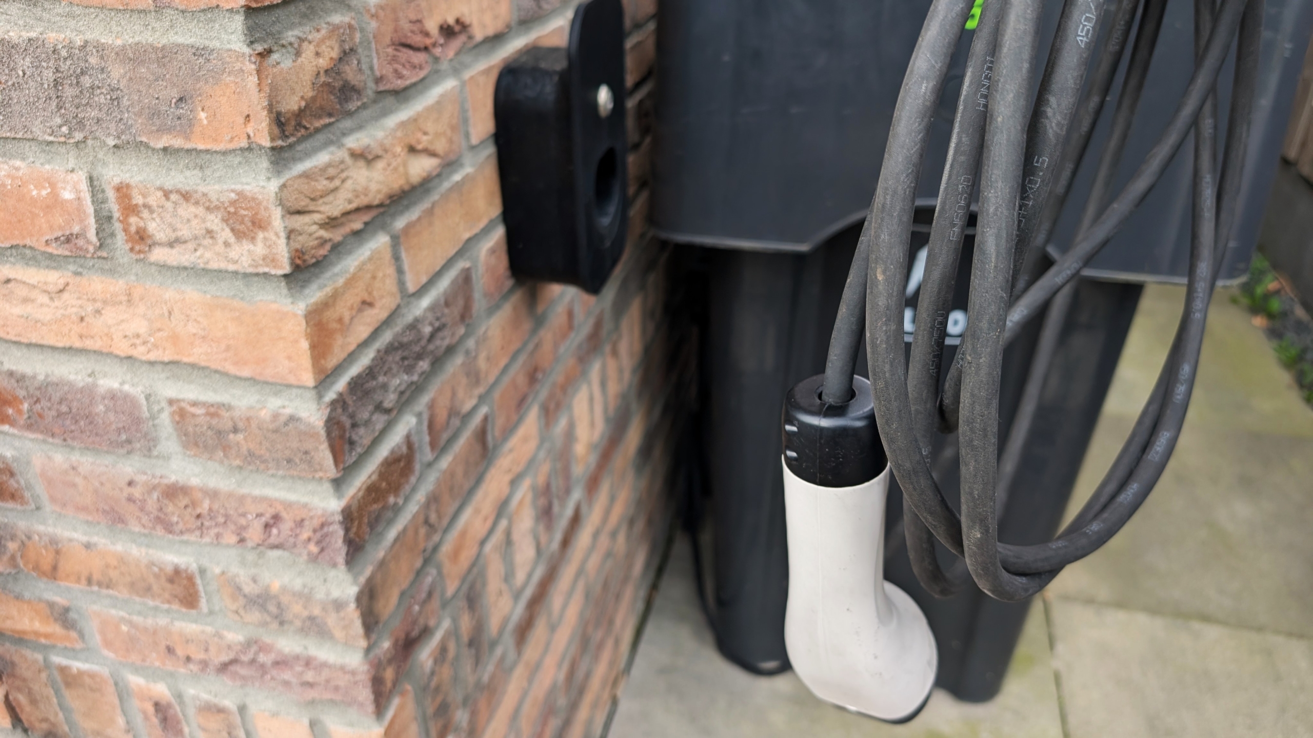 Een elektrische laadkabel hangt naast een bakstenen muur, met een laadstation op de achtergrond.