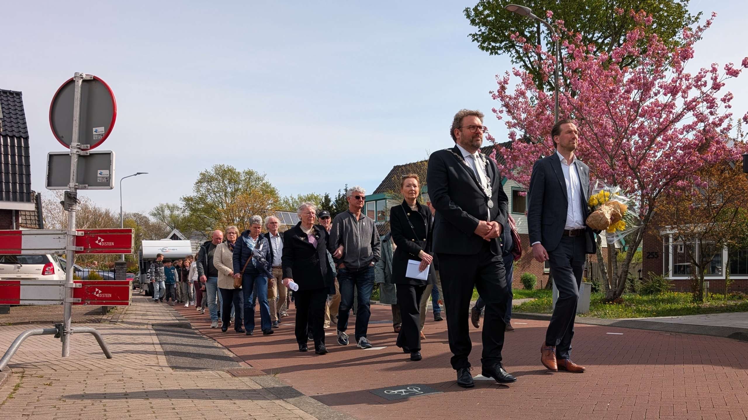 Mensen lopen in een stoet over een straat onder bloeiende bomen, met een persoon die bloemen draagt aan de voorkant.