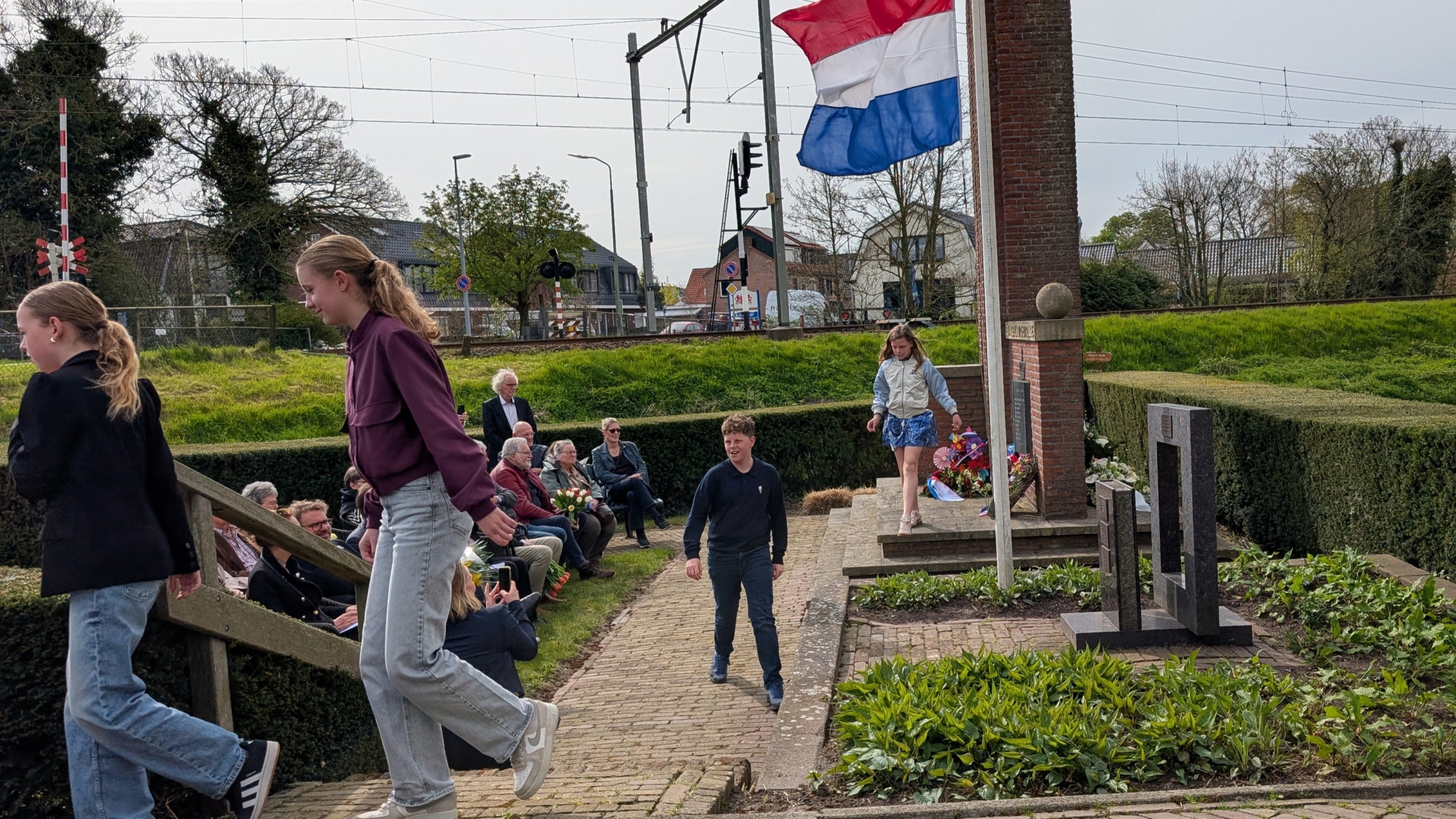 Mensen bij een gedenkteken met een Nederlandse vlag; sommigen zitten, anderen lopen voorbij.