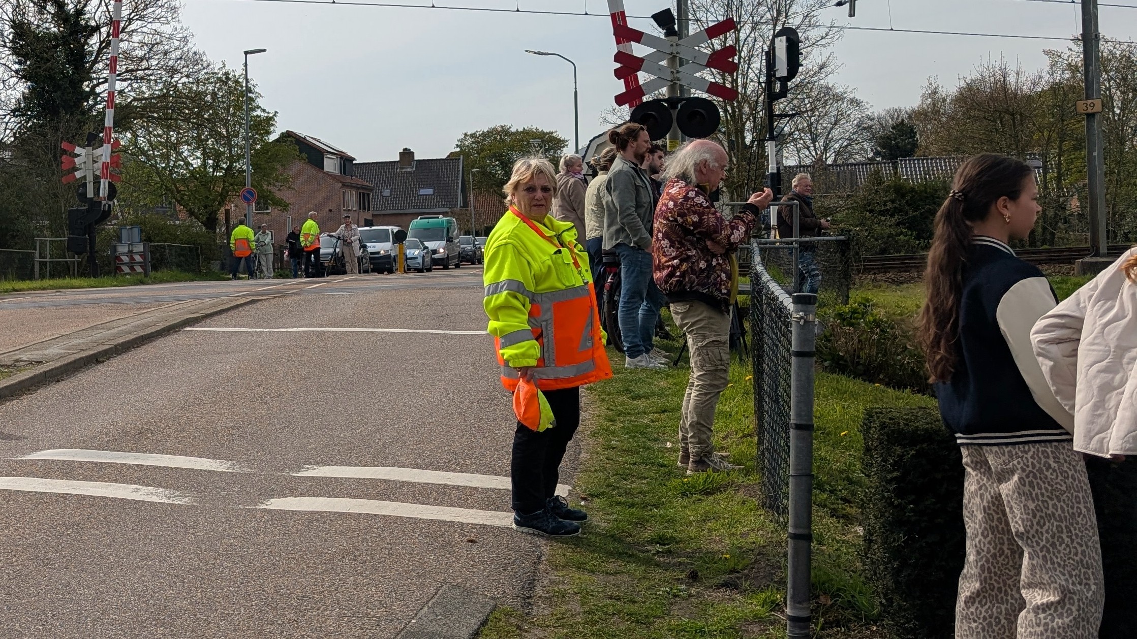 Mensen staan te wachten bij een spoorwegovergang; een persoon draagt een opvallend geel en oranje veiligheidshesje.