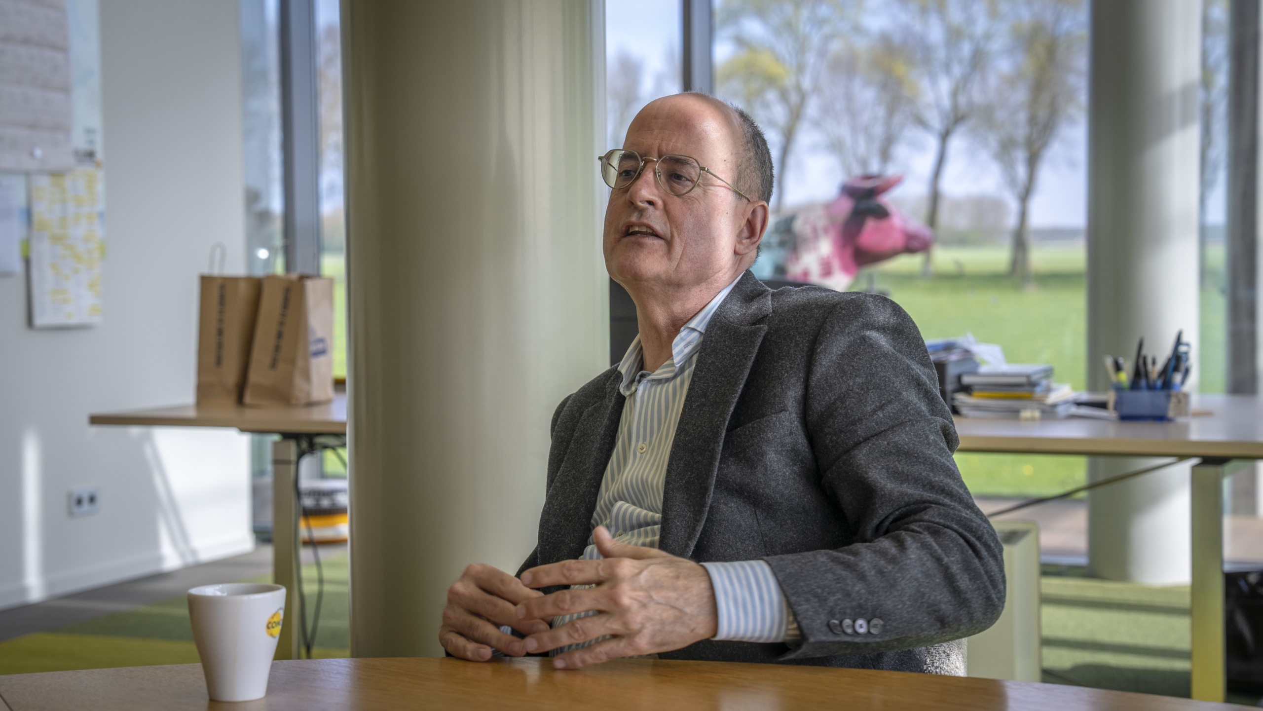 Man met bril en grijs jasje zit aan een tafel in een kantoor met papieren tasjes en een kartonnen beker.