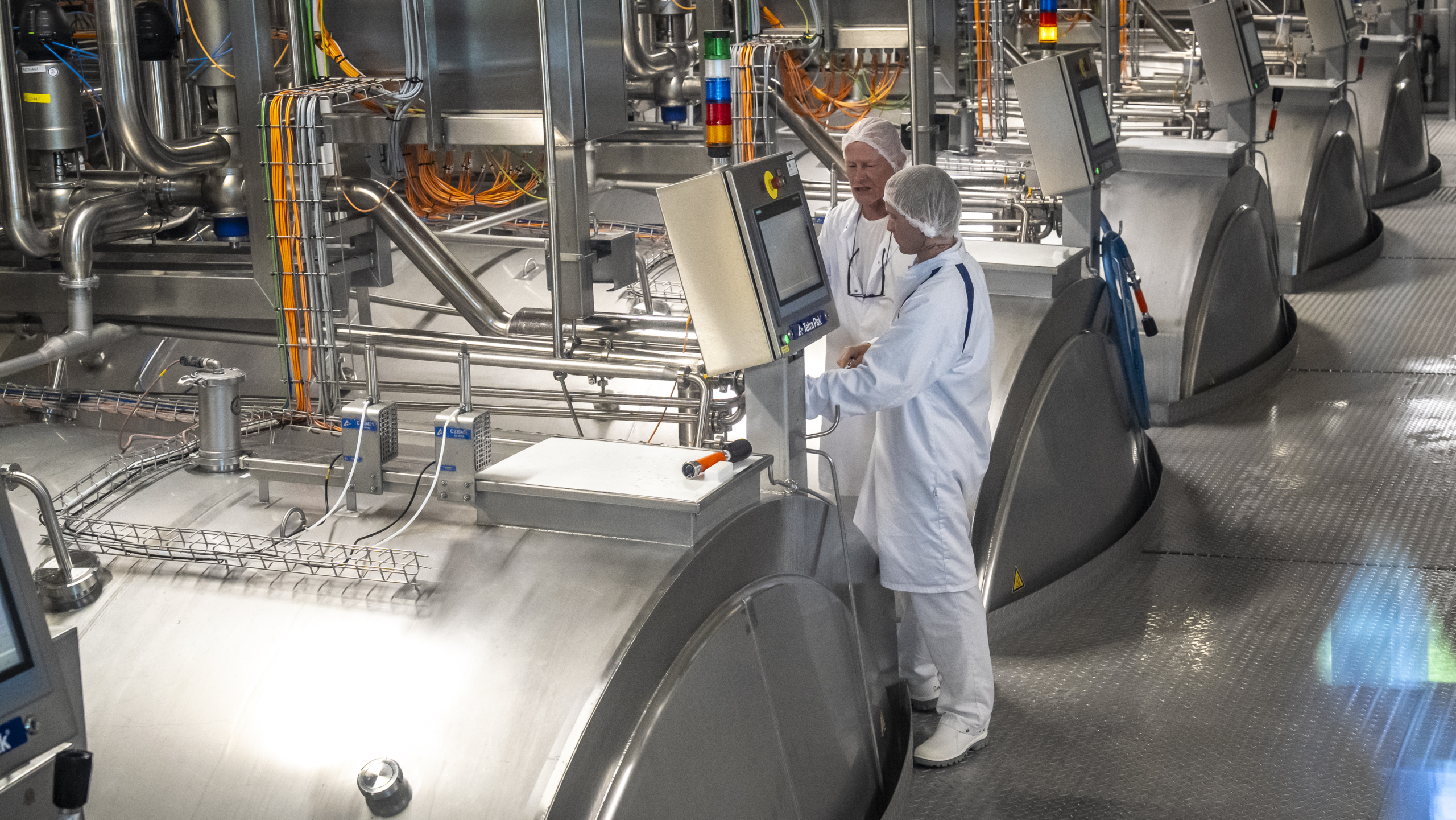 Twee mensen in witte beschermende kleding bedienen een monitor in een industriële fabriek met metalen apparaten en buizen.