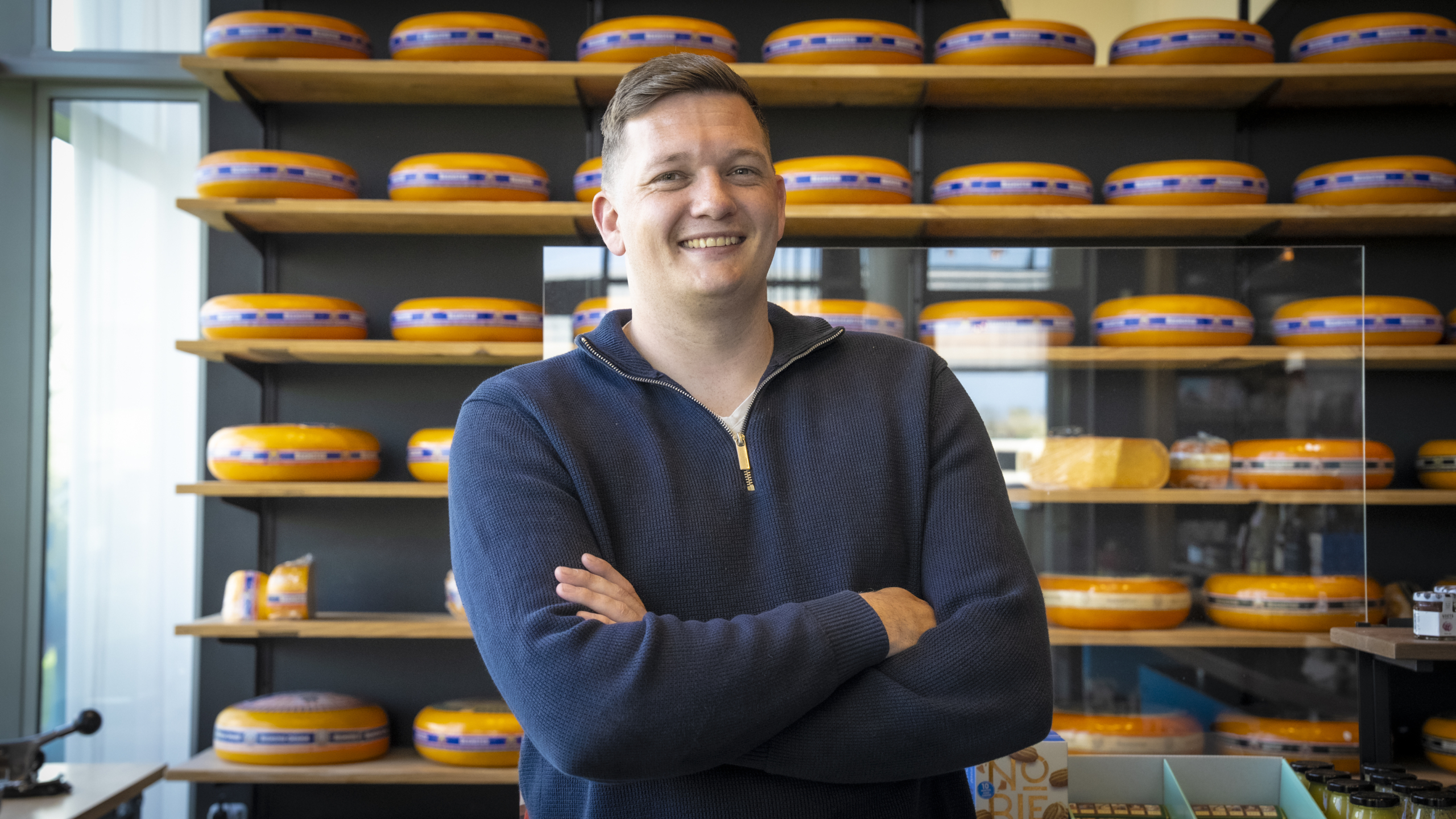 Man in een donkerblauwe trui staat glimlachend voor planken met ronde kazen op de achtergrond.
