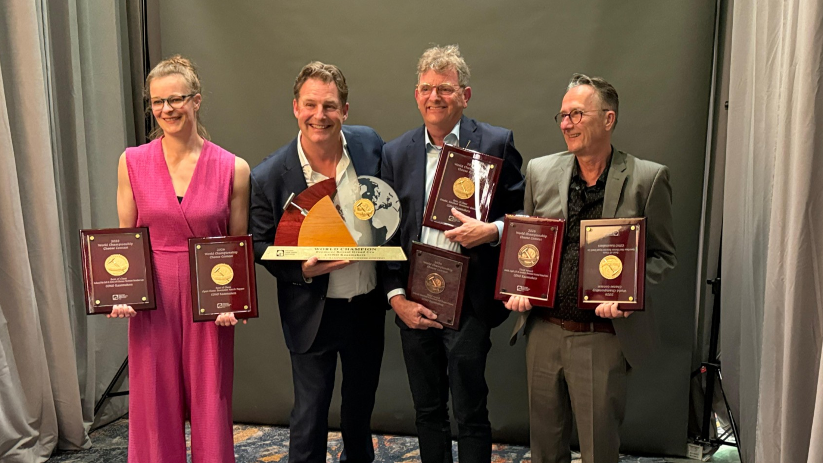 Vier mensen poseren met trots en houden trofeeën en onderscheidingen vast in een formele setting.