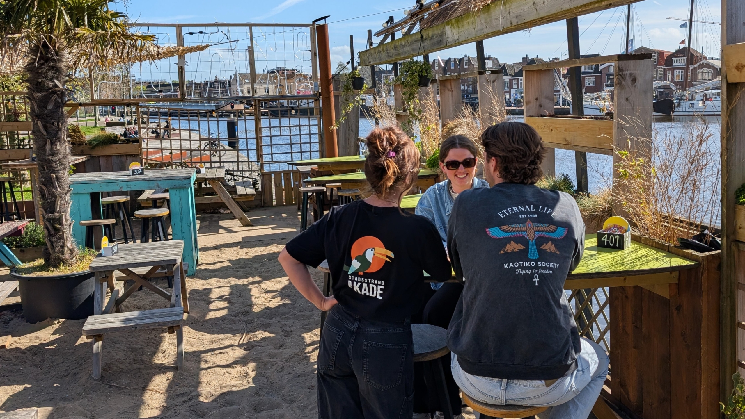 Drie mensen zitten aan een tafel in een strandbar nabij een haven, omgeven door houten meubels en planten.