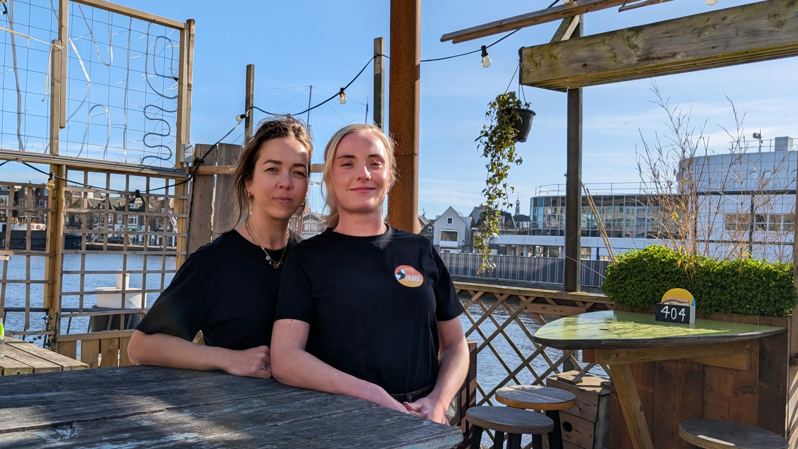 Twee vrouwen in zwarte shirts bij een houten tafel op een terras met uitzicht op een rivier en gebouwen op de achtergrond.