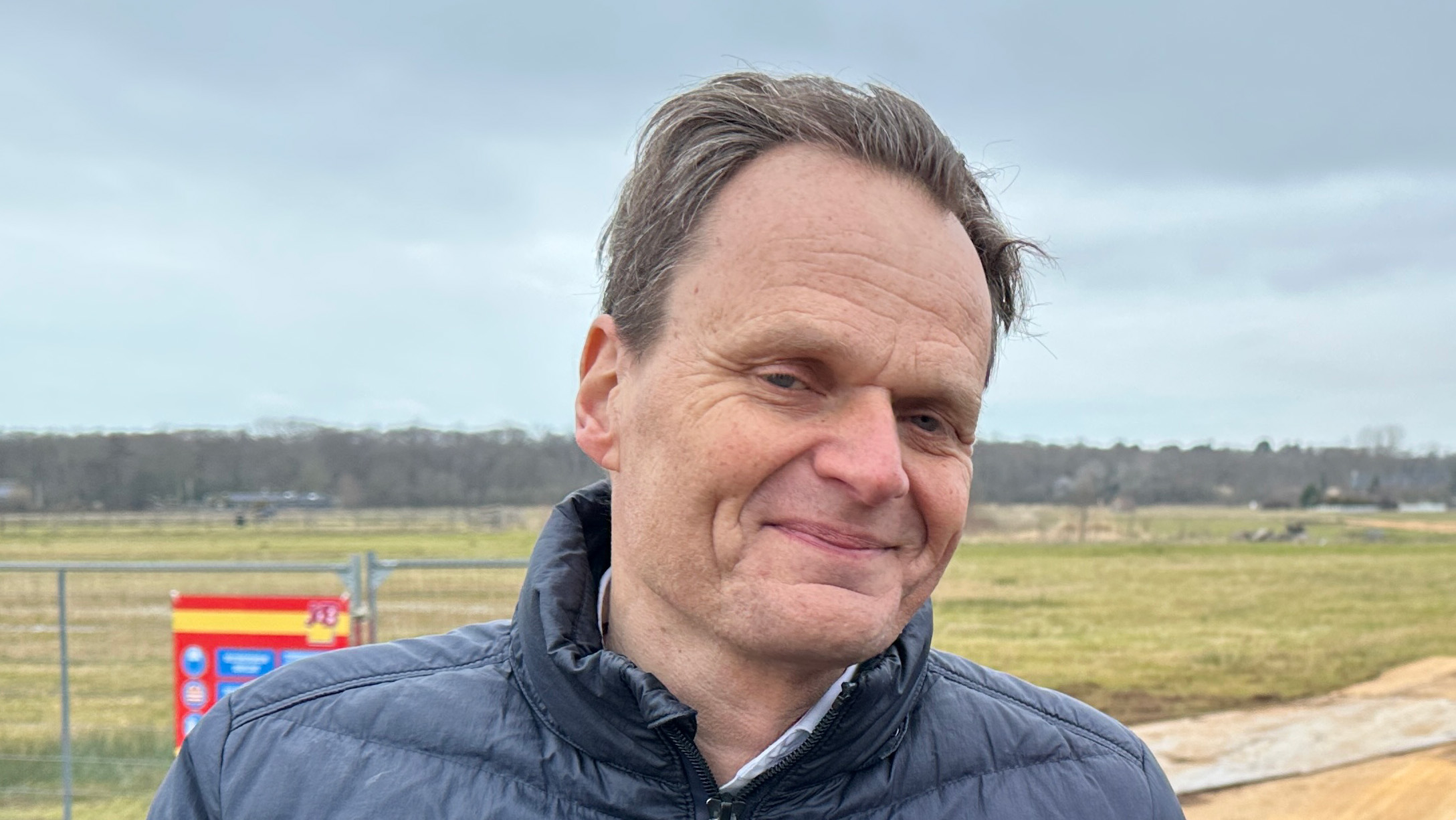 Man in een grijze jas glimlacht buiten op een open veld onder een bewolkte hemel.