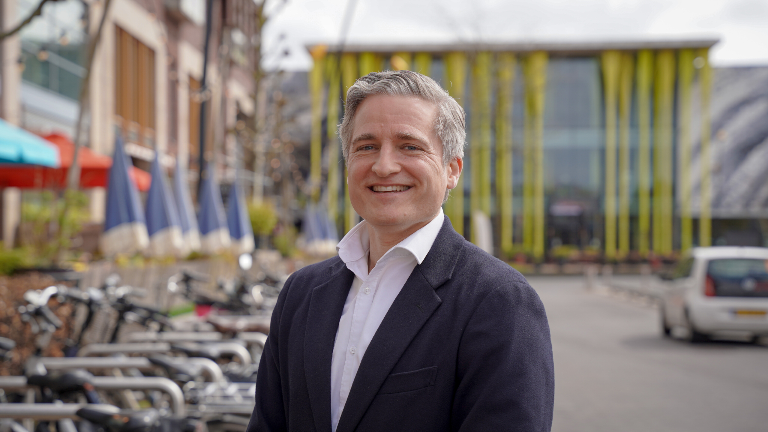 Man in een donkerblauwe blazer glimlacht buiten, met fietsen en een modern gebouw op de achtergrond.