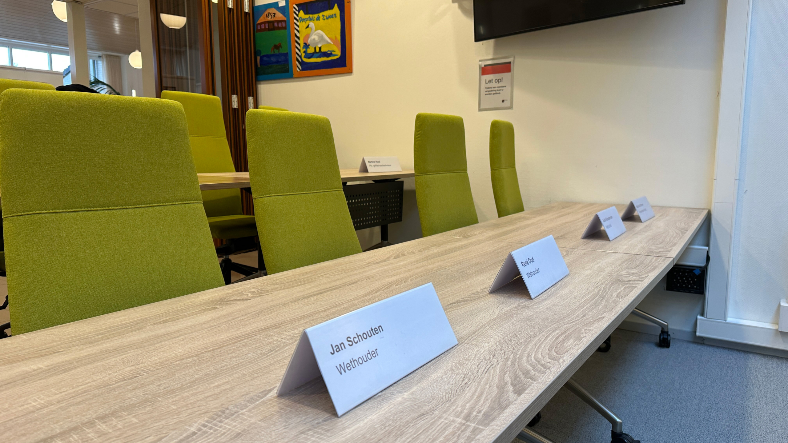 Vergaderruimte met groene stoelen en naamkaartjes op een houten tafel, waarop onder andere "Jan Schouten, Wethouder" staat.