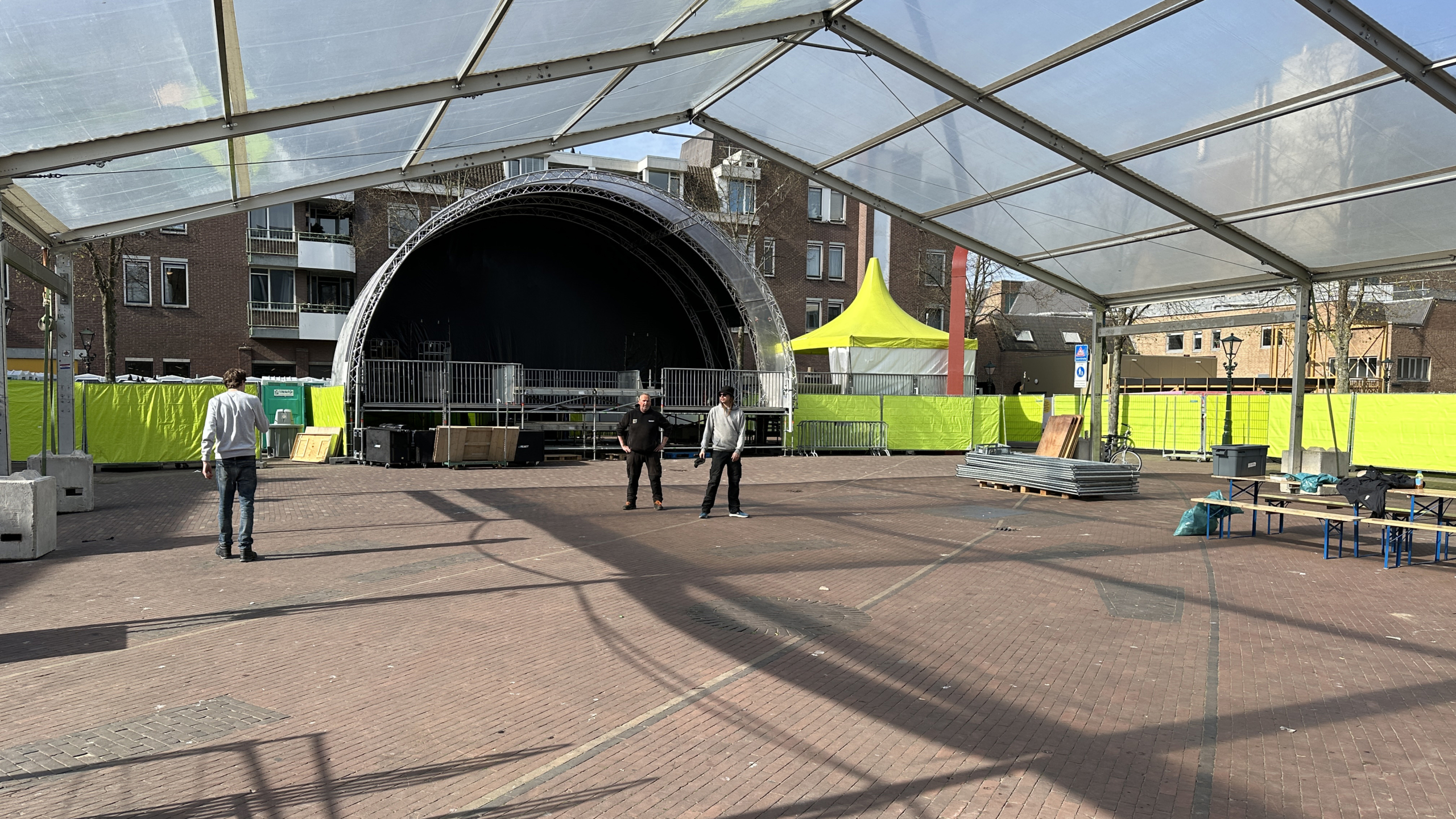 Twee mannen staan op een leeg plein voor een grote openluchtpodium, omgeven door fluoriscerende gele hekken en een geel tent.