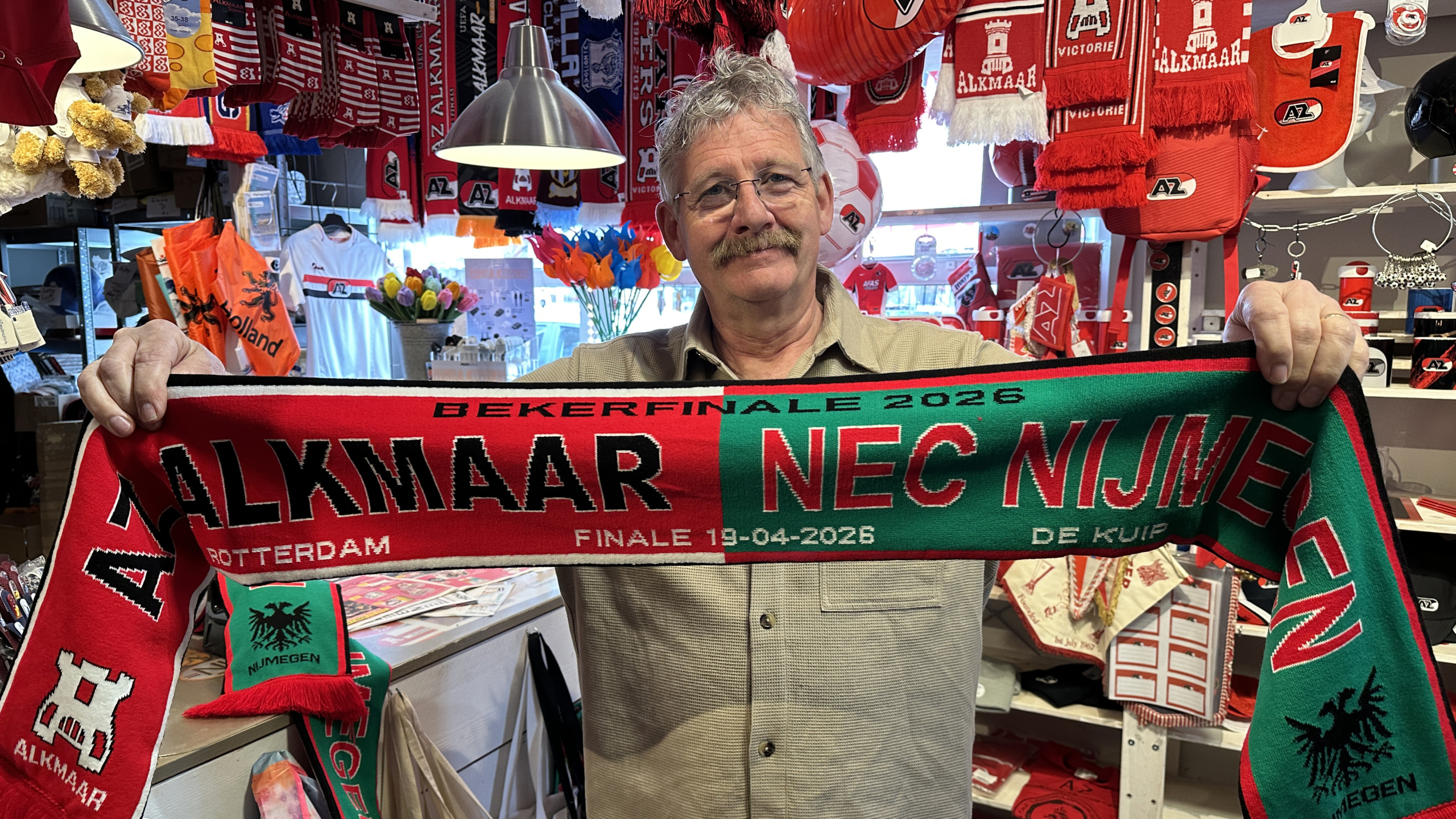 Man met een sjaal voor de bekerfinale tussen Alkmaar en NEC in 2026, omringd door sportmerchandising.