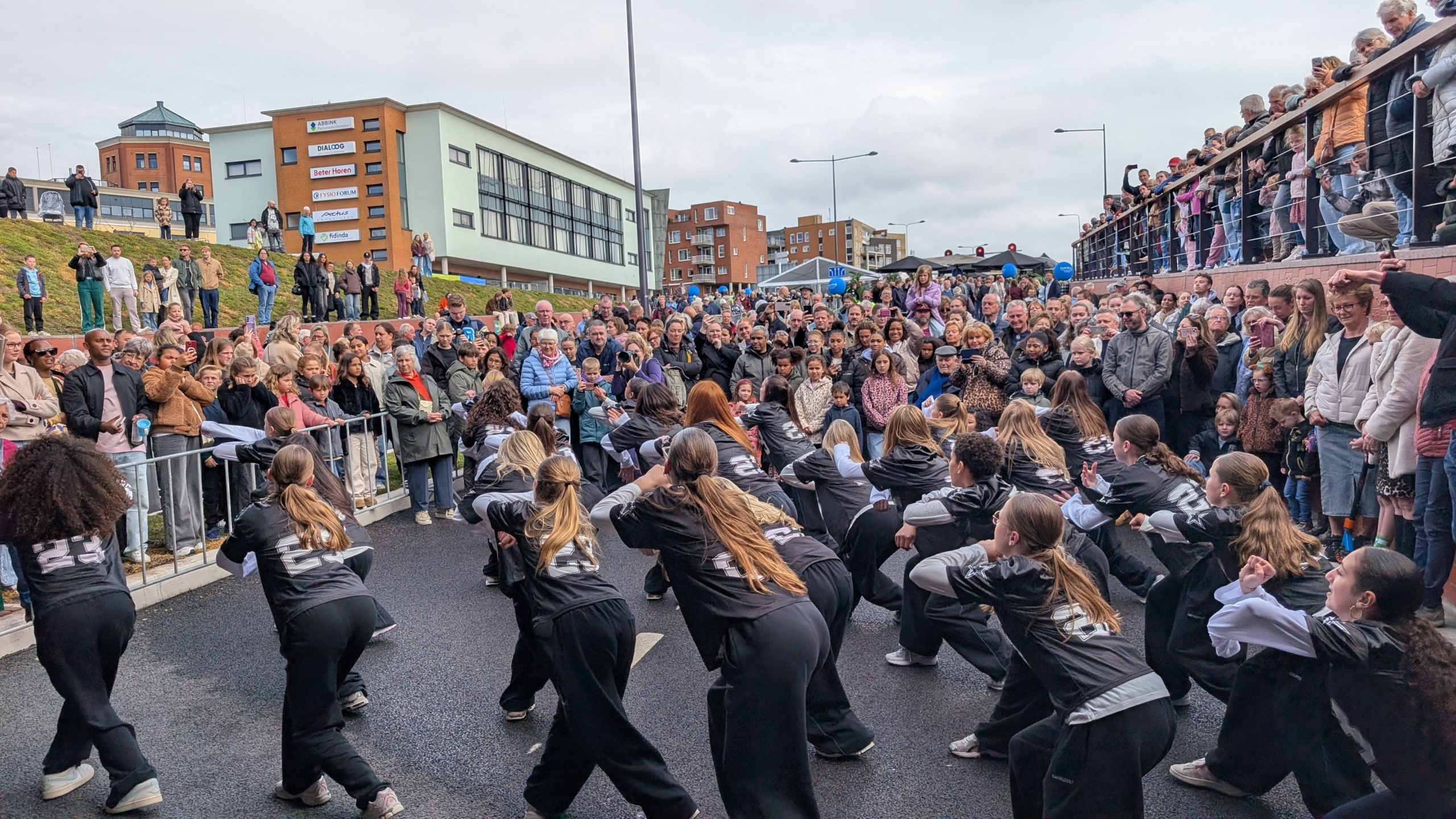 Een dansgroep voert een optreden op voor een grote, opgewekte menigte op een straat, met een gebouw en toeschouwers op een helling op de achtergrond.