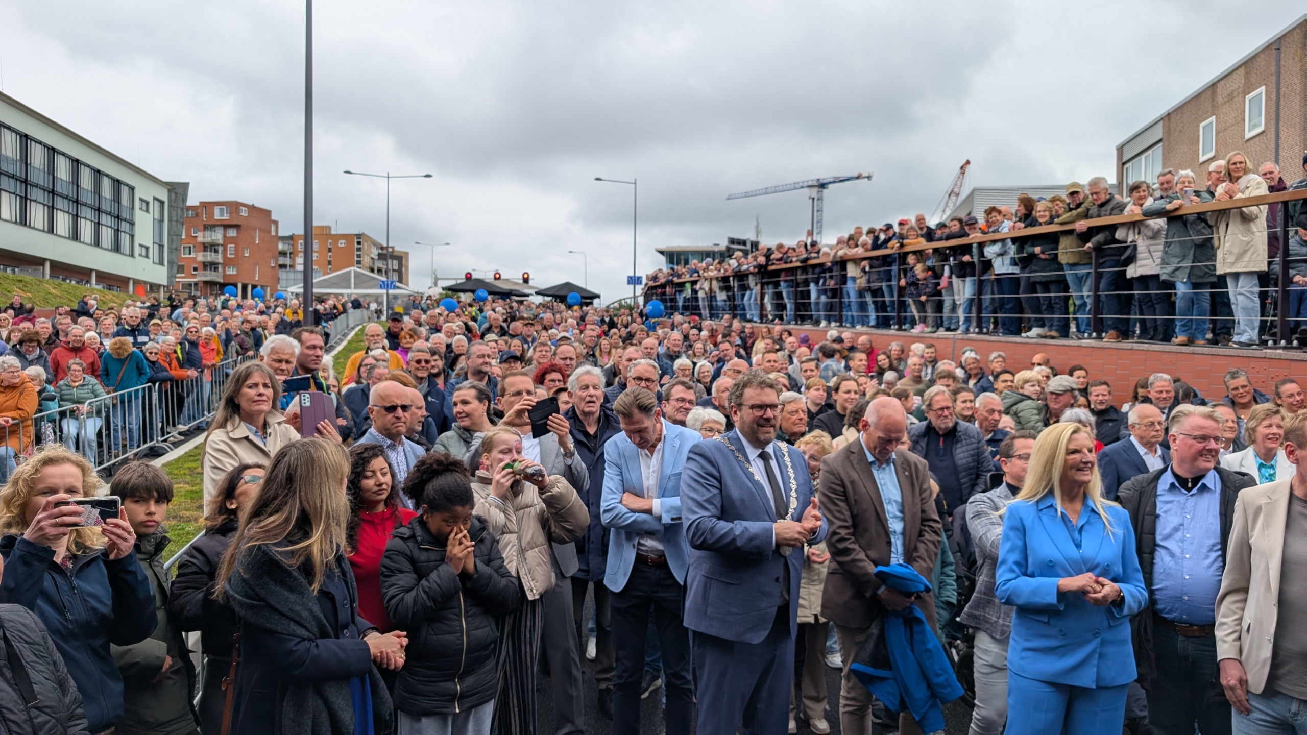 Een grote menigte mensen staat buiten, enkele nemen foto's tijdens een openbare bijeenkomst.