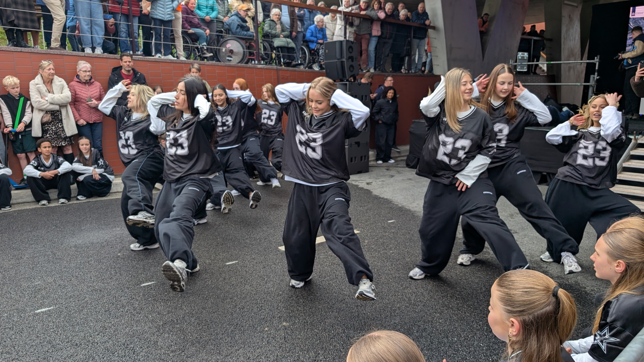 Groep meisjes in zwarte sportoutfits voert een dansroutine op, met een publiek dat toekijkt.