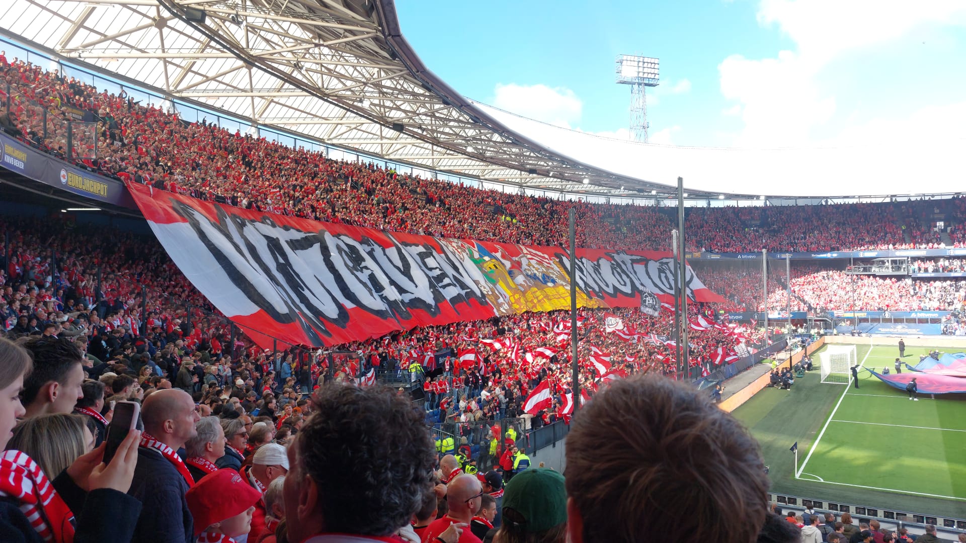 Vol stadion gevuld met supporters, grote rode en witte spandoek met tekst wordt omhoog gehouden boven menigte.