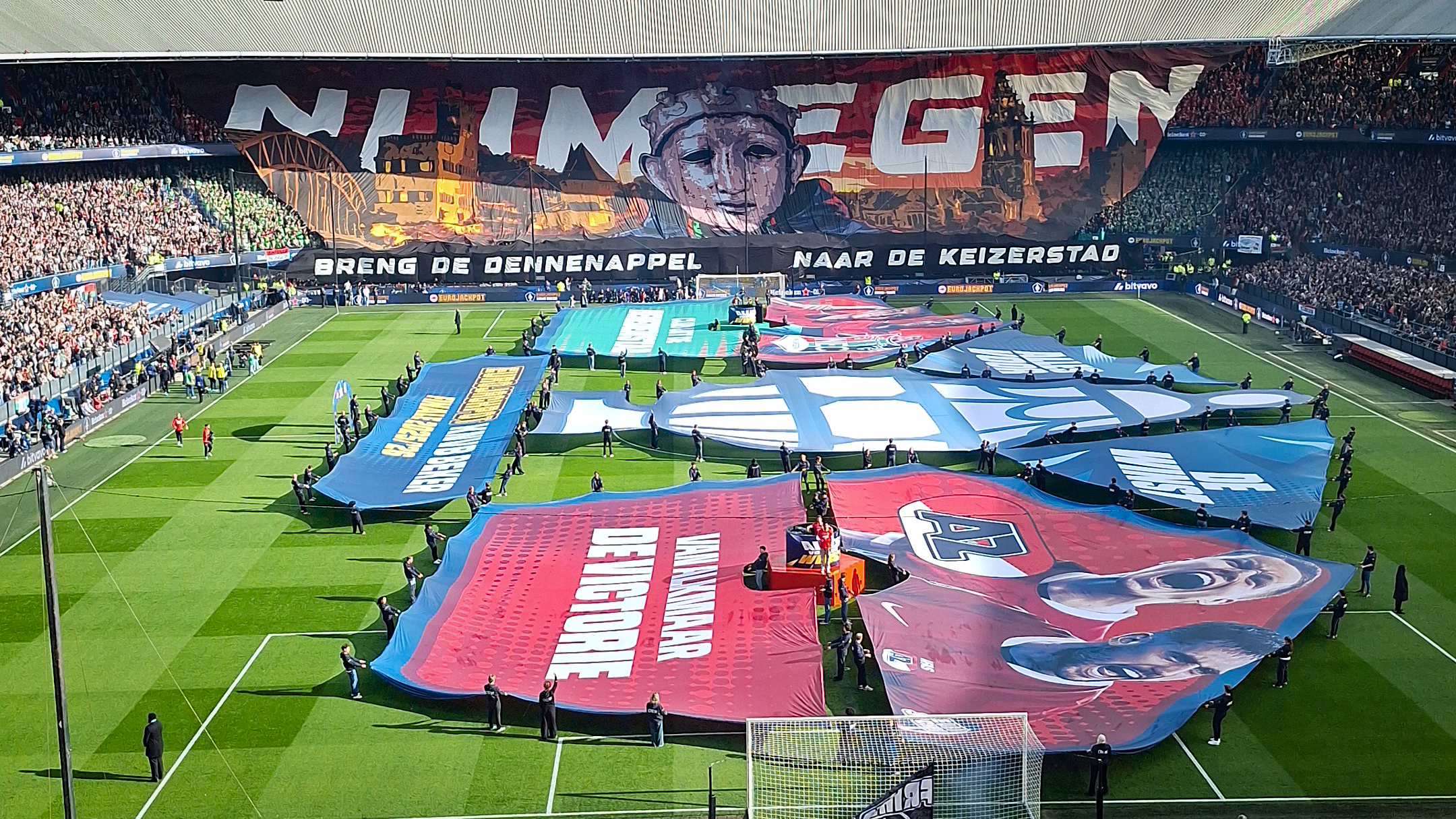 Voetbalstadion met groot spandoek in publiek met de tekst "Nijmegen" en grote doeken op het veld gedragen door mensen.