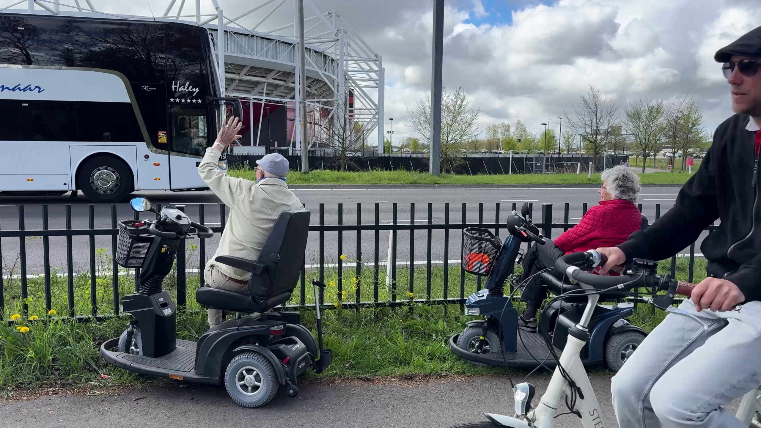 Een man in een scootmobiel zwaait naar een voorbijrijdende bus, naast een vrouw in een rode jas en een fietser op de voorgrond.