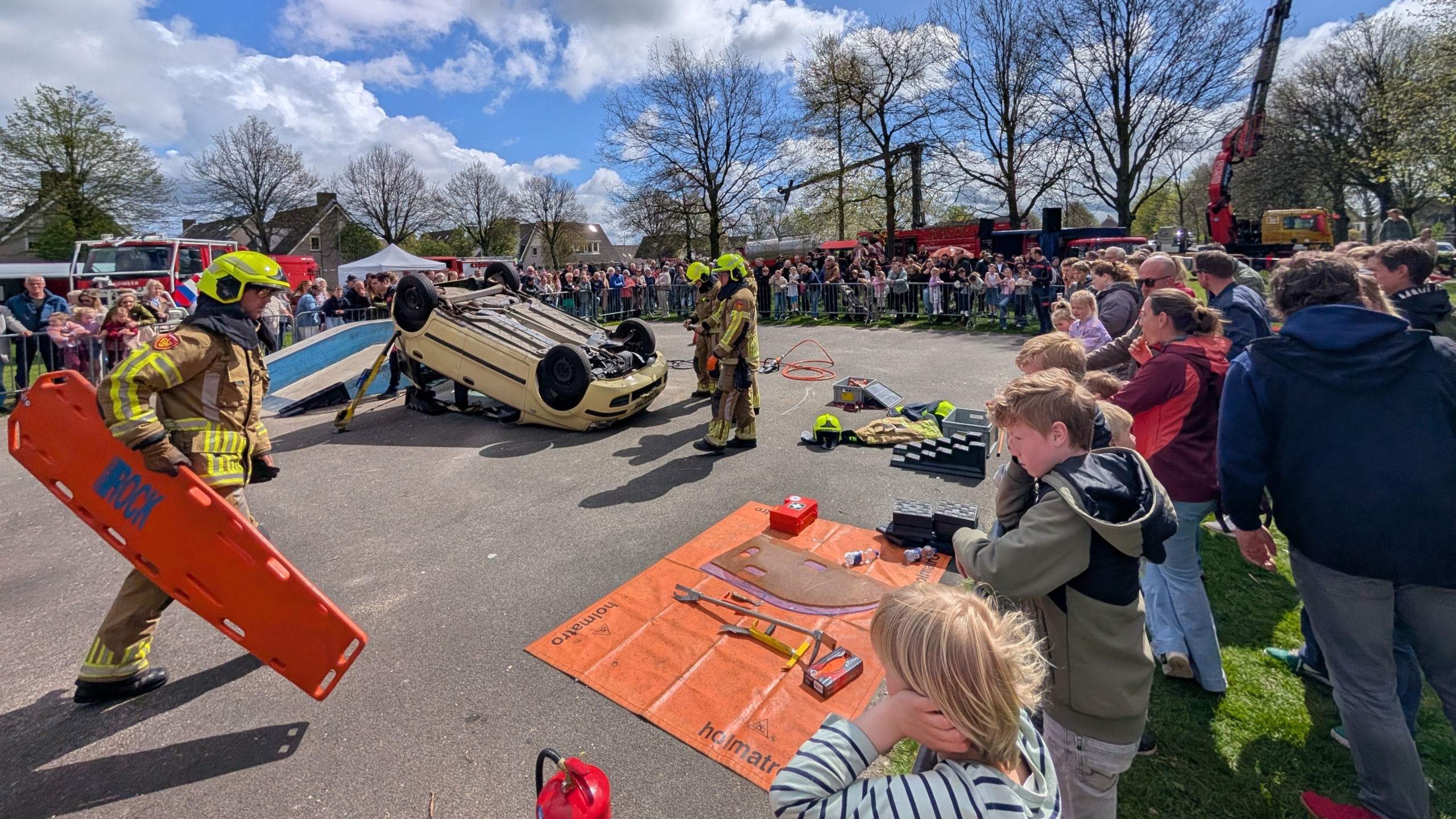 Brandweer voert een demonstratie uit met een omgekeerde auto, omringd door toeschouwers op een zonnige dag.