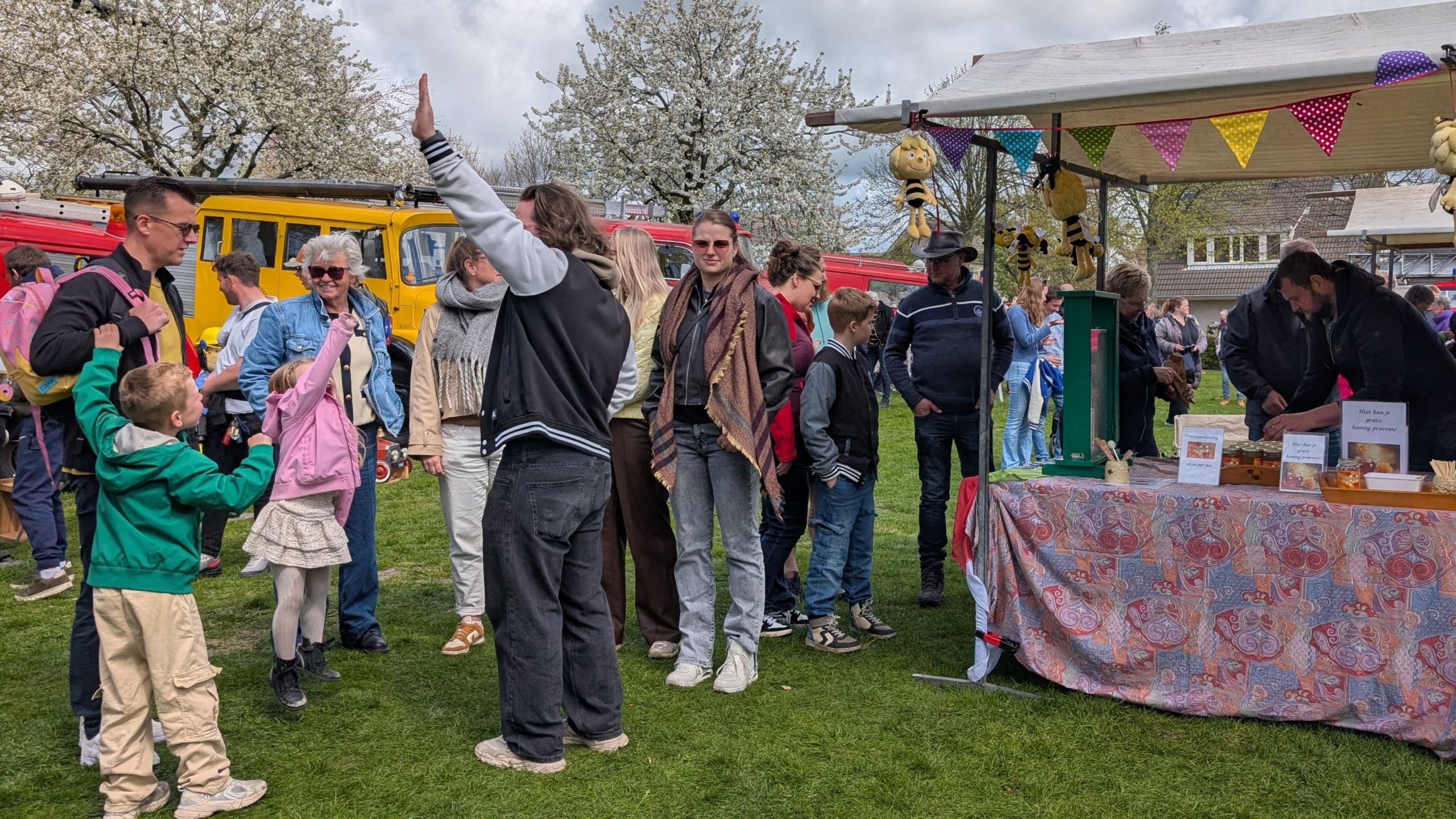 Mensen genieten van een drukbezochte markt met kraampjes, kleurrijke vlaggetjes en bloeiende bomen op de achtergrond.