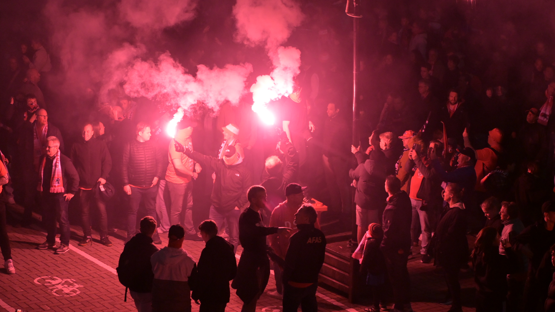 Mensen in een menigte houden fakkels vast die roze rook afgeven, terwijl anderen toekijken.