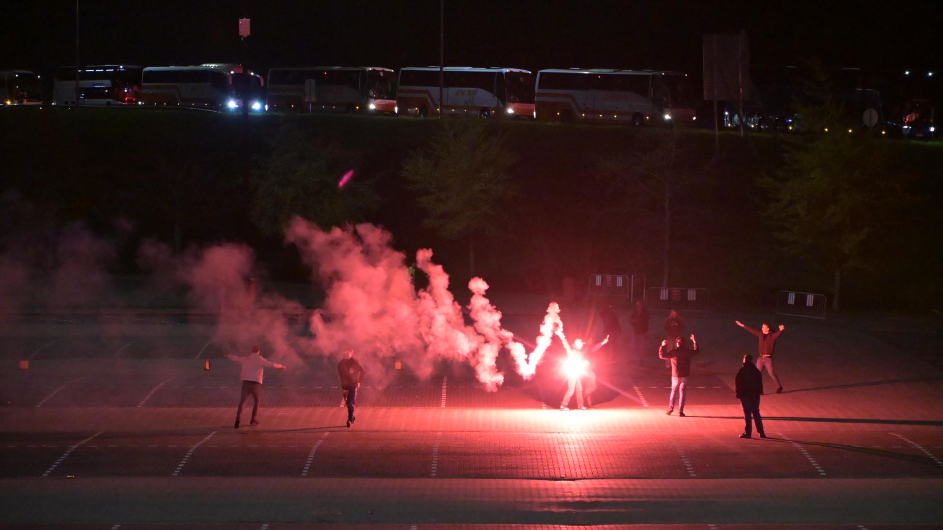 Mensen spelen met fakkels die rood licht en rook verspreiden op een parkeerplaats in de avond, omgeven door bussen.