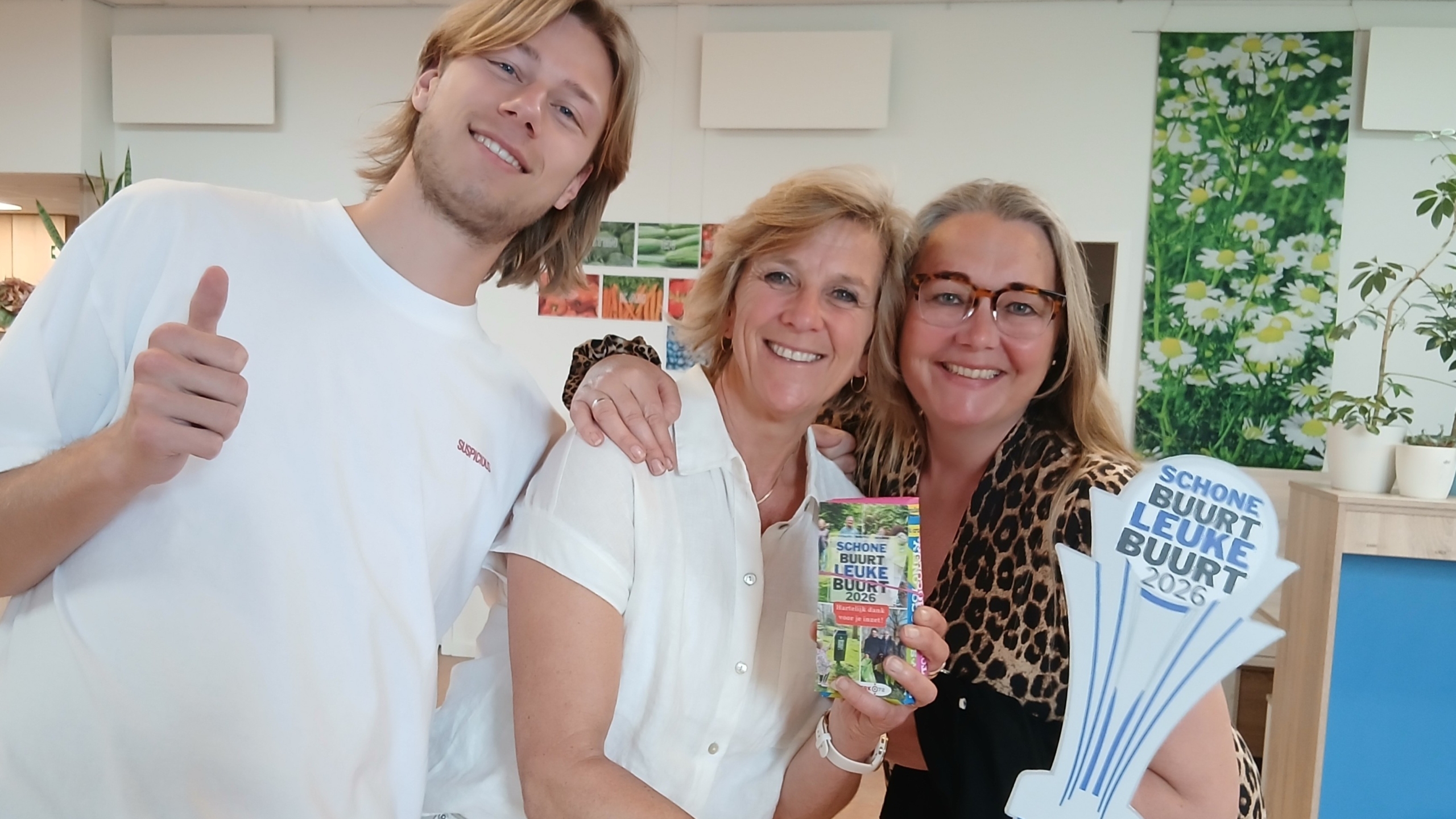Drie mensen poseren vrolijk voor de camera, een van hen houdt een object met tekst "Schone Buurt Leuke Buurt 2026" vast.