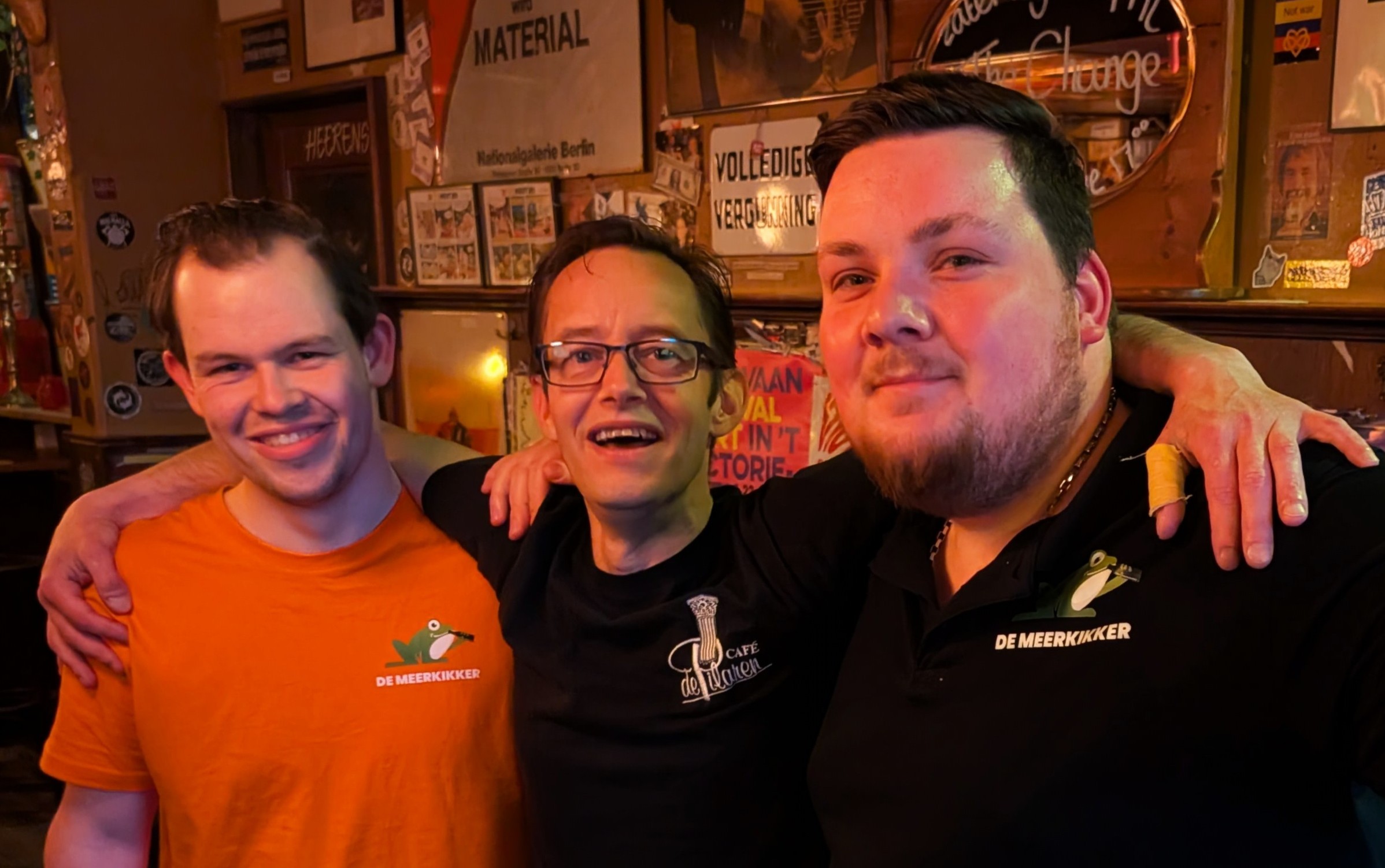 Drie mannen glimlachen in een bar, met de armen om elkaar heen. Op de achtergrond zijn barposters en decoraties te zien.