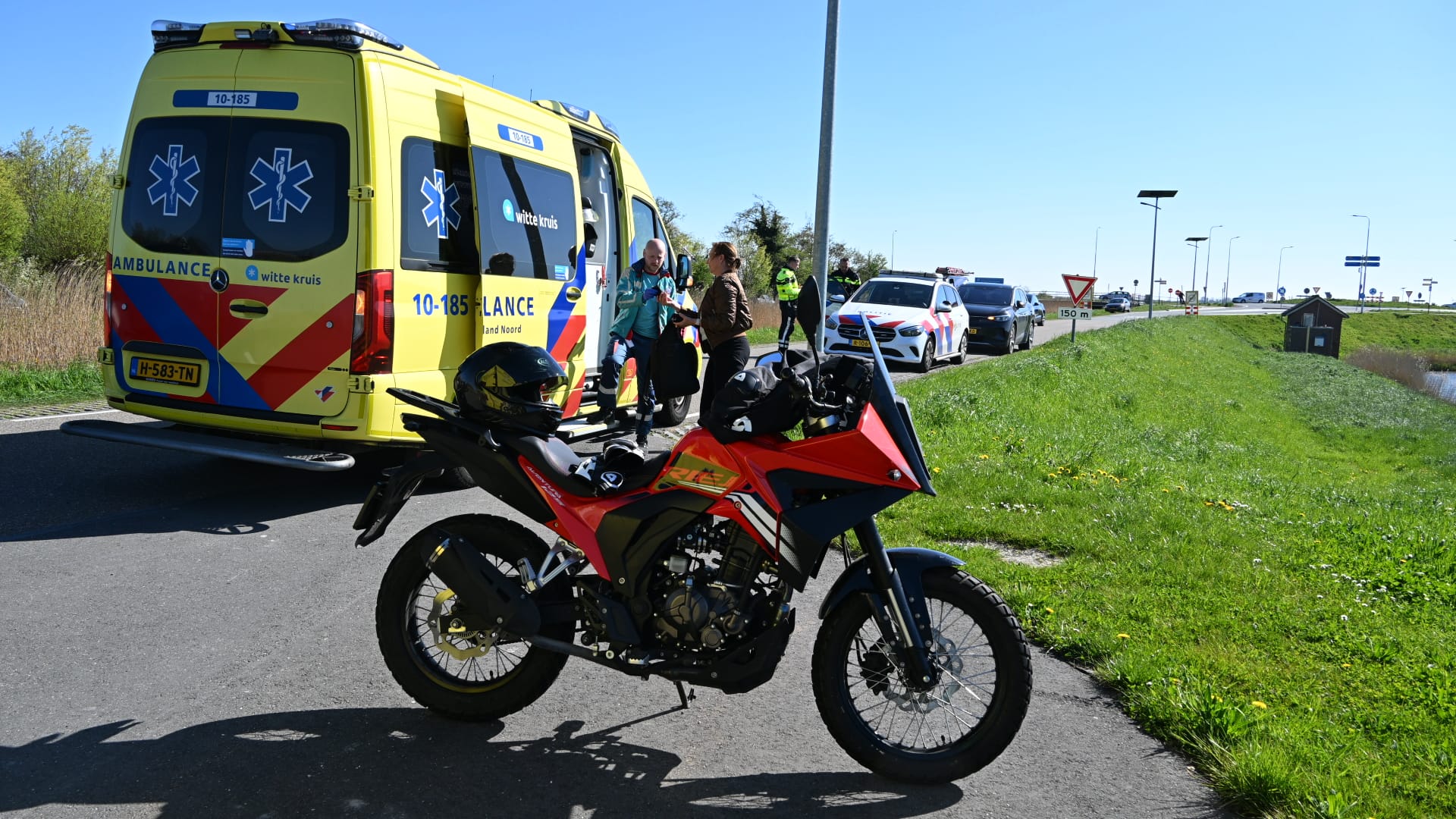 Ambulance en politieauto staan naast een motorfiets op een zonnige dag, met mensen die bij de ambulance staan.