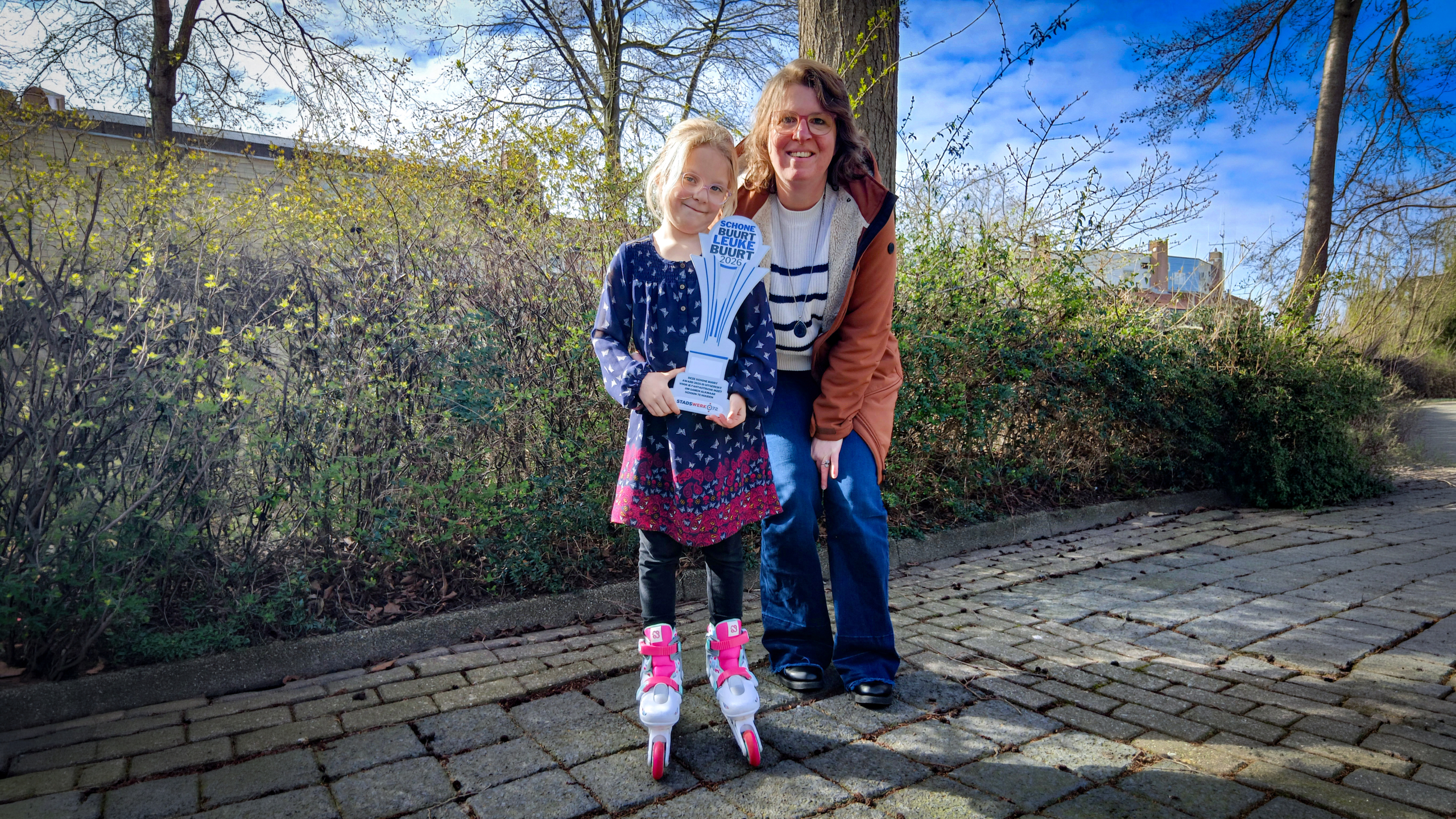 Een jong meisje op skeelers houdt een trofee vast, naast haar staat een glimlachende volwassen vrouw op een zonnige dag buiten.