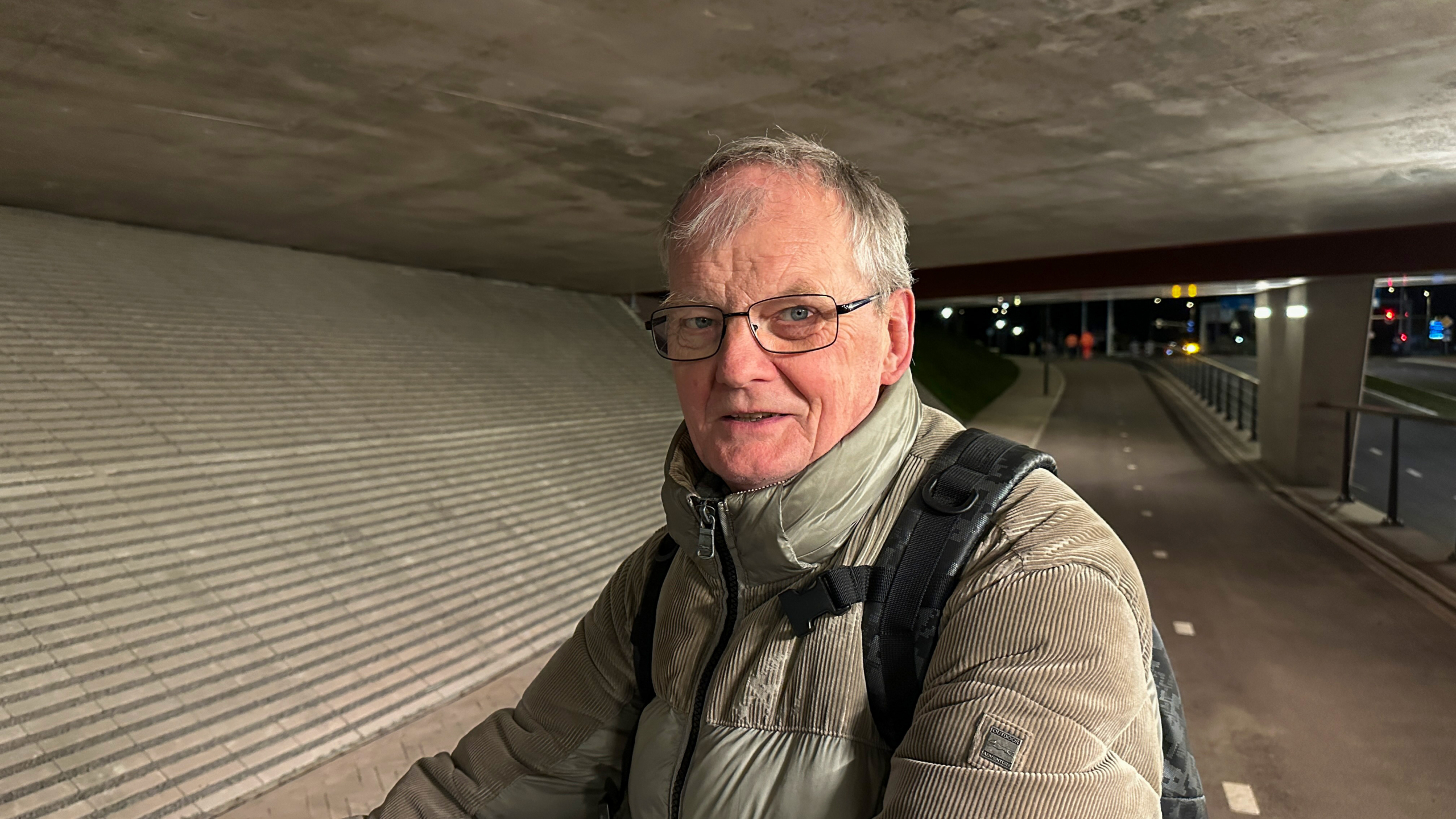 Man met een bril en grijze jas staat onder een viaduct, met verlichting en fietspaden zichtbaar in de achtergrond.