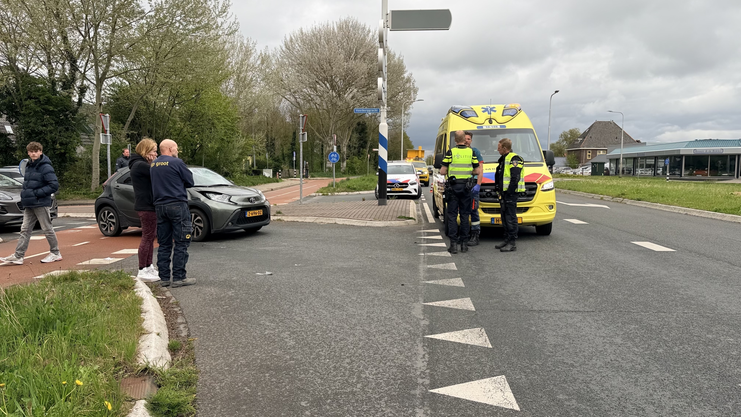 Politieagenten en een ambulance op een kruispunt, met enkele mensen bij een auto betrokken bij een incident.