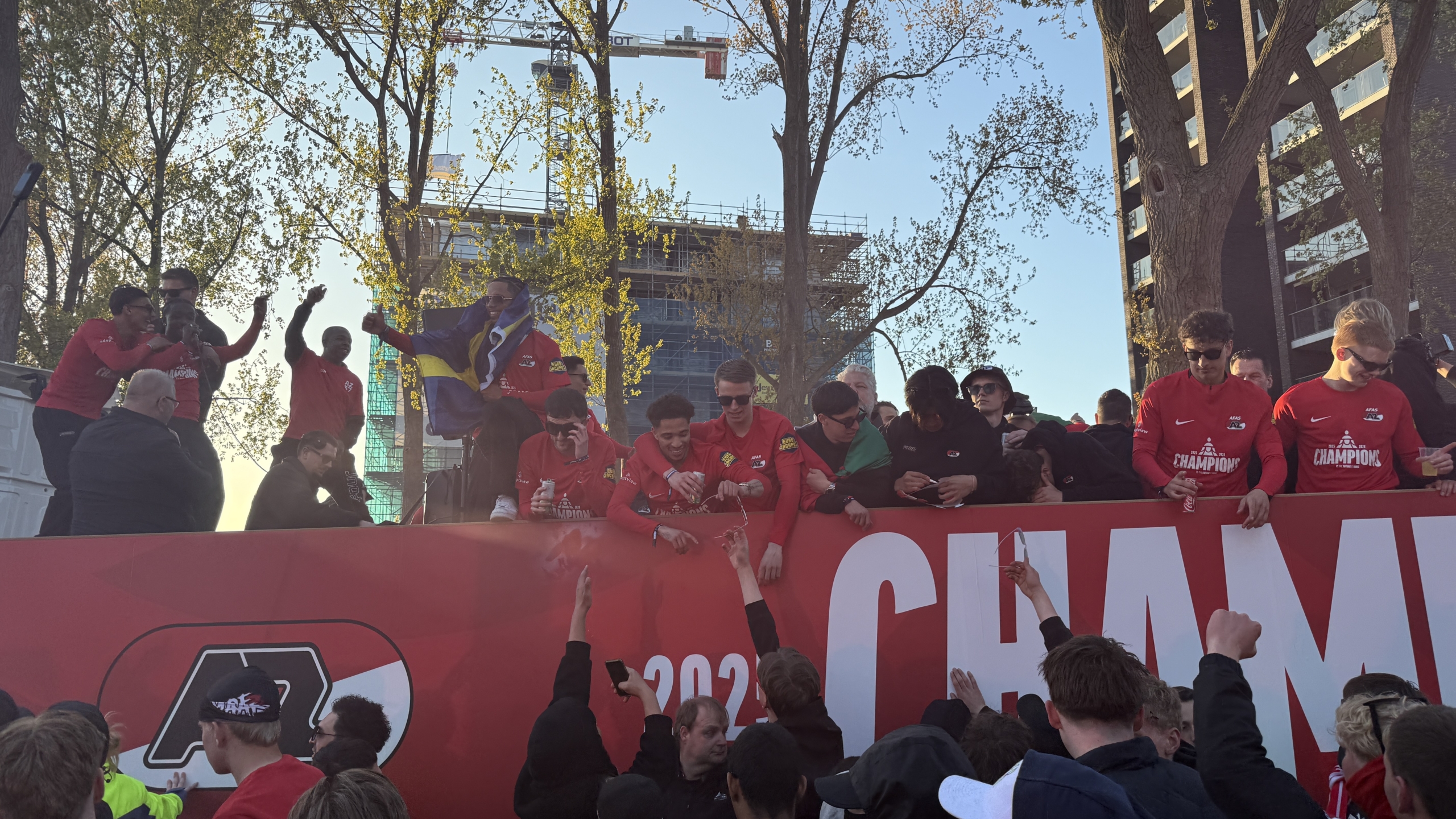 Mensen in rode shirts vieren feest op een tribune met de tekst "Champion", omringd door fans die juichen.